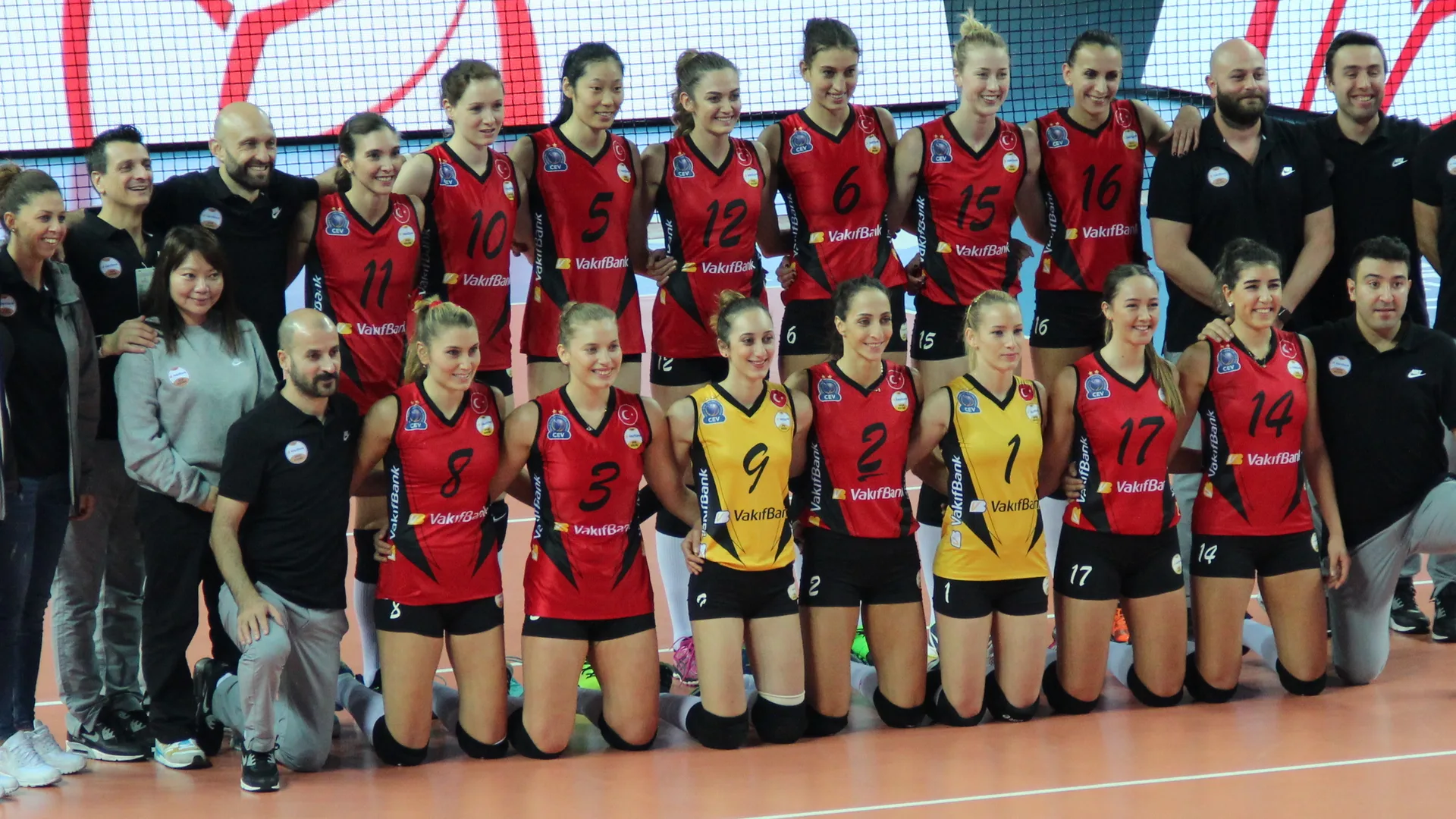 Kırmızı ve sarı formalı kadın voleybolcular teknik ekiple sahada toplu poz veriyor