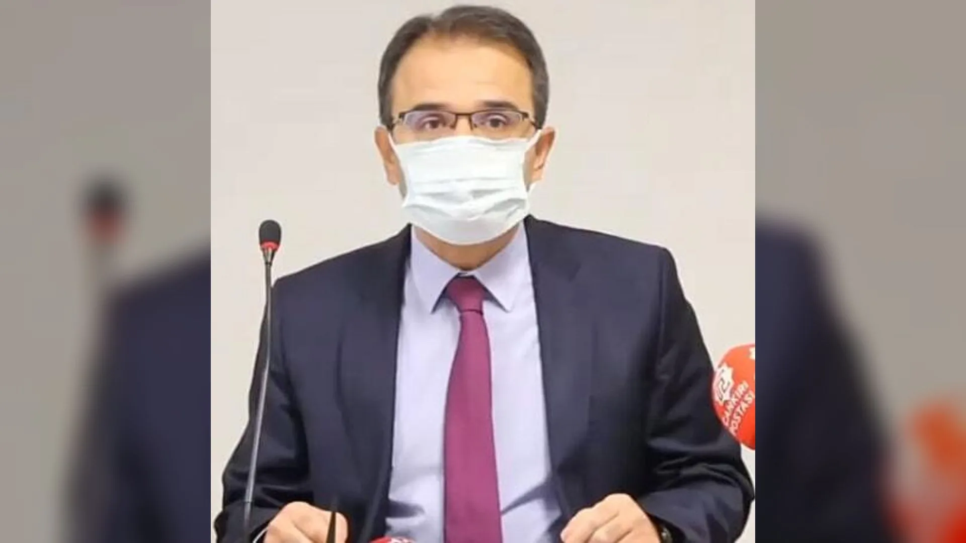 Maske ve gözlük takan, lacivert takım elbiseli ve mor kravatlı bir adam mikrofon önünde konuşuyor