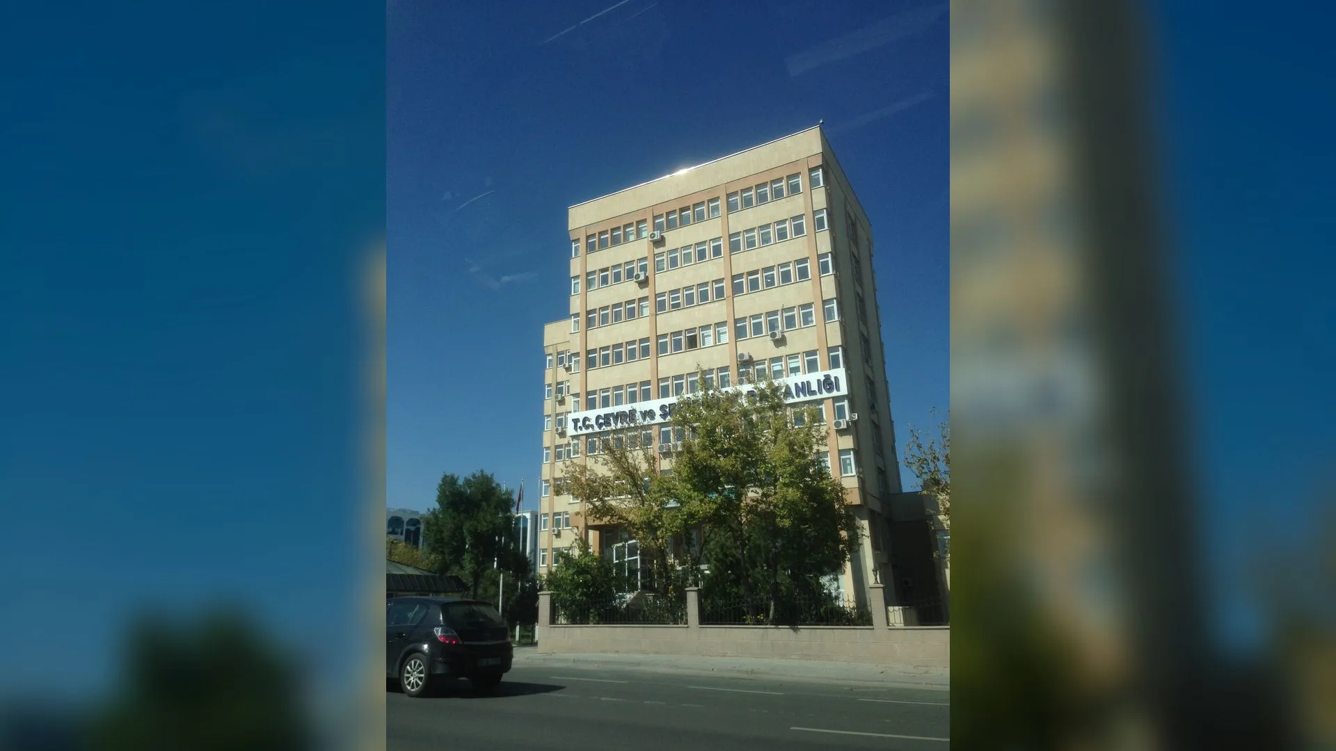 Açık mavi gökyüzü altında, ağaçların arkasında tabelalı, çok katlı bej renkli bir bakanlık binası