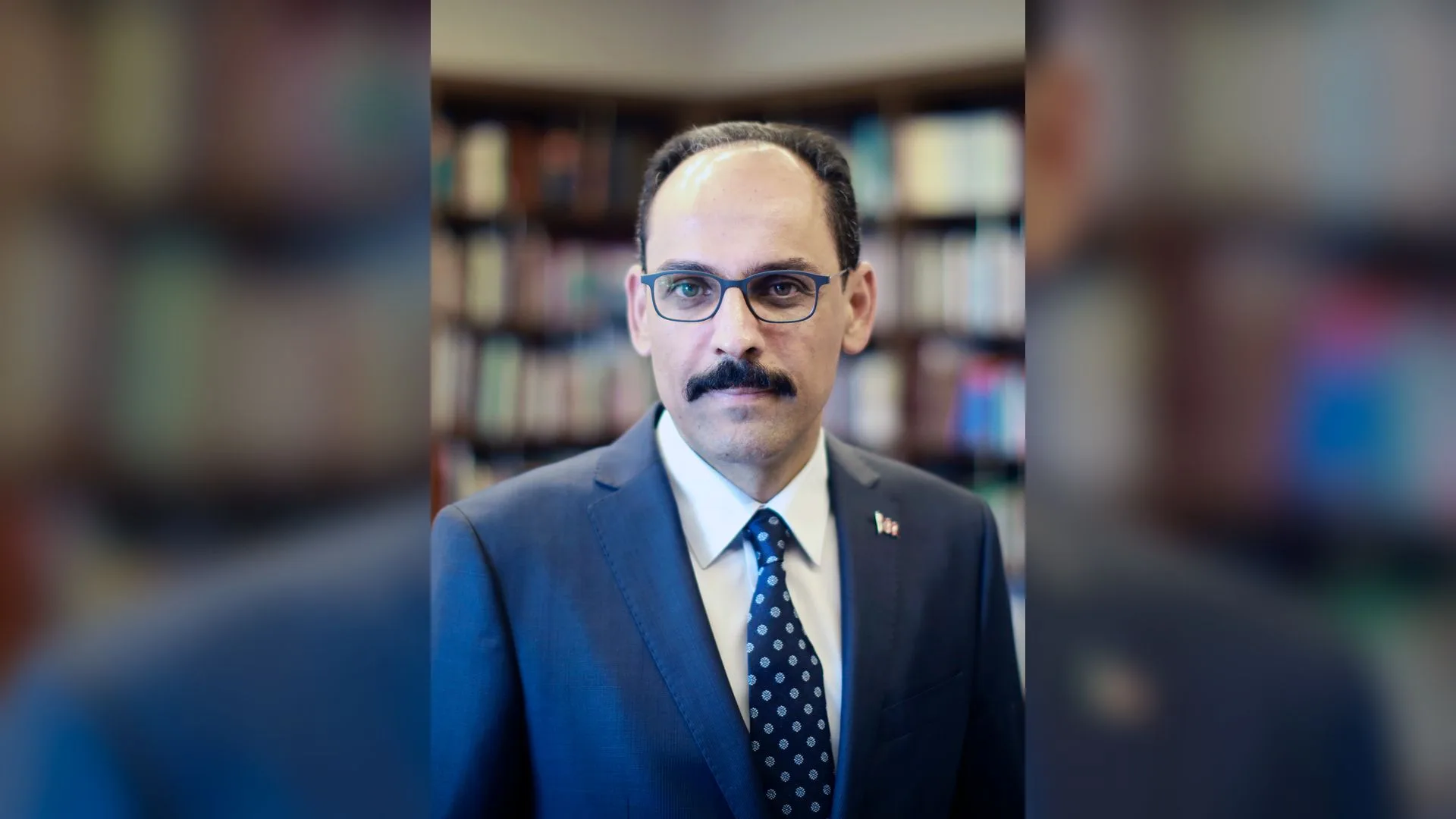 Kitaplık önünde gözlüklü, bıyıklı, lacivert takım elbiseli ve puantiyeli kravatlı bir adam