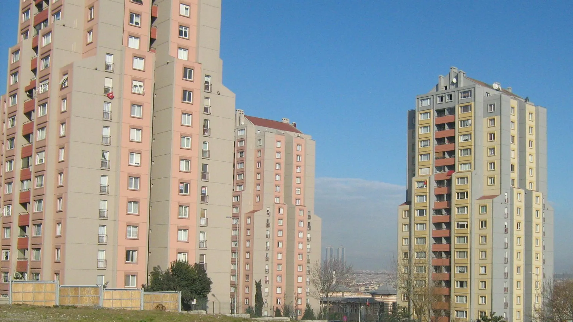 Açık mavi gökyüzü altında, boş bir arazinin arkasında yükselen pembe ve sarı renkli yüksek konut blokları