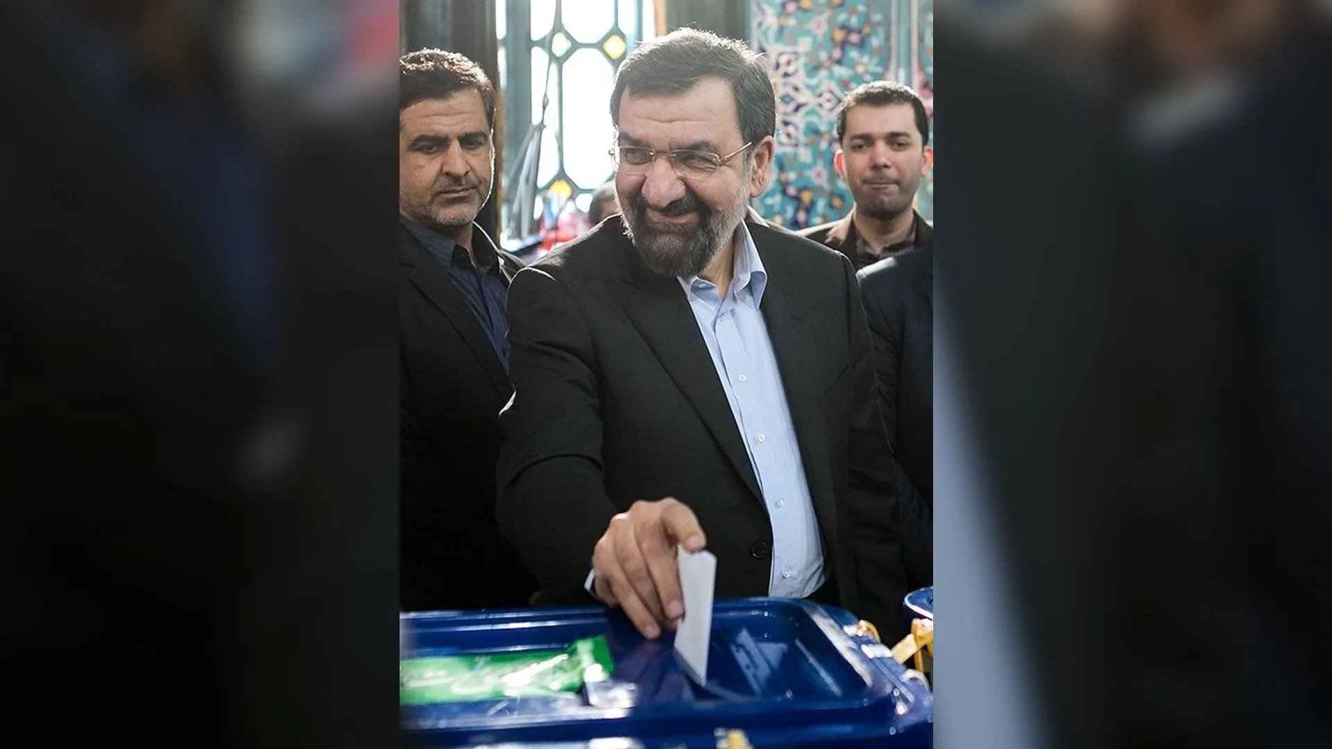 Gözlüklü, sakallı takım elbiseli bir adam gülümseyerek mavi sandığa oy pusulası atıyor