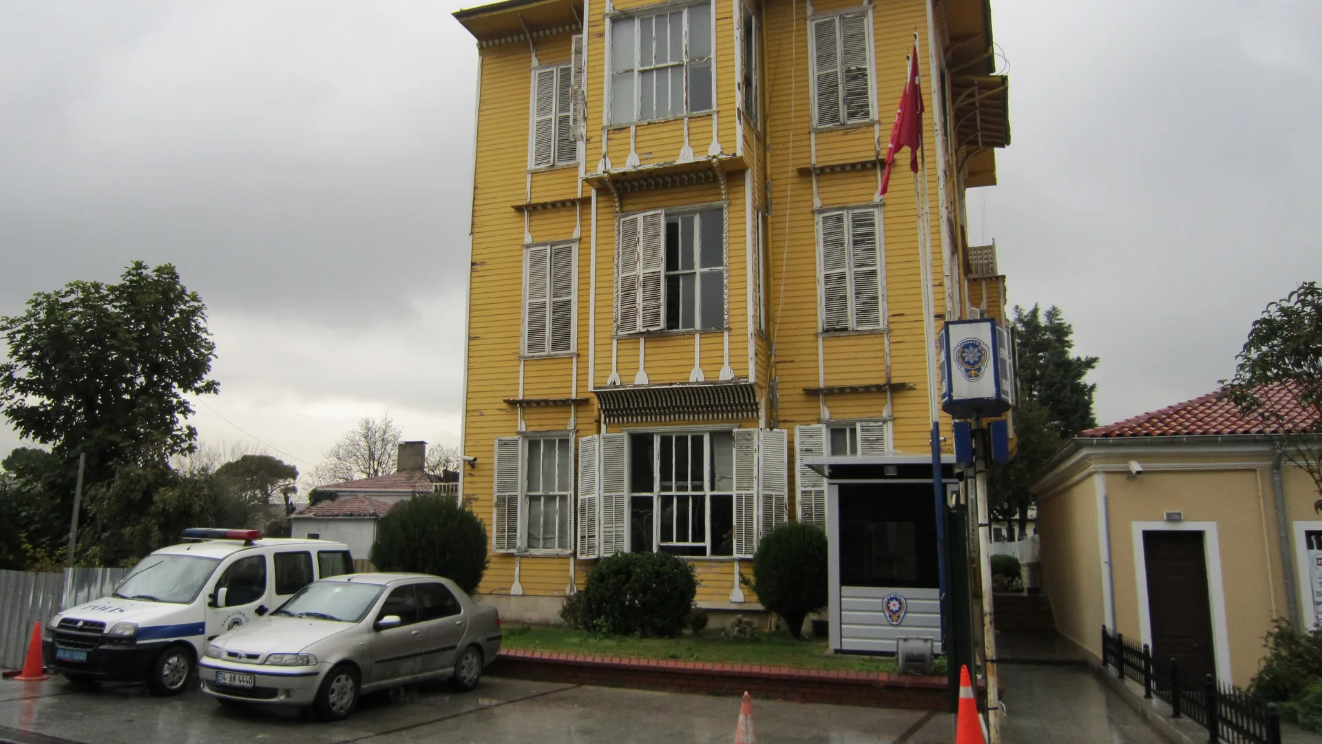 Bulutlu havada, Türk bayrağı ve polis tabelası bulunan sarı ahşap, üç katlı tarihi bina