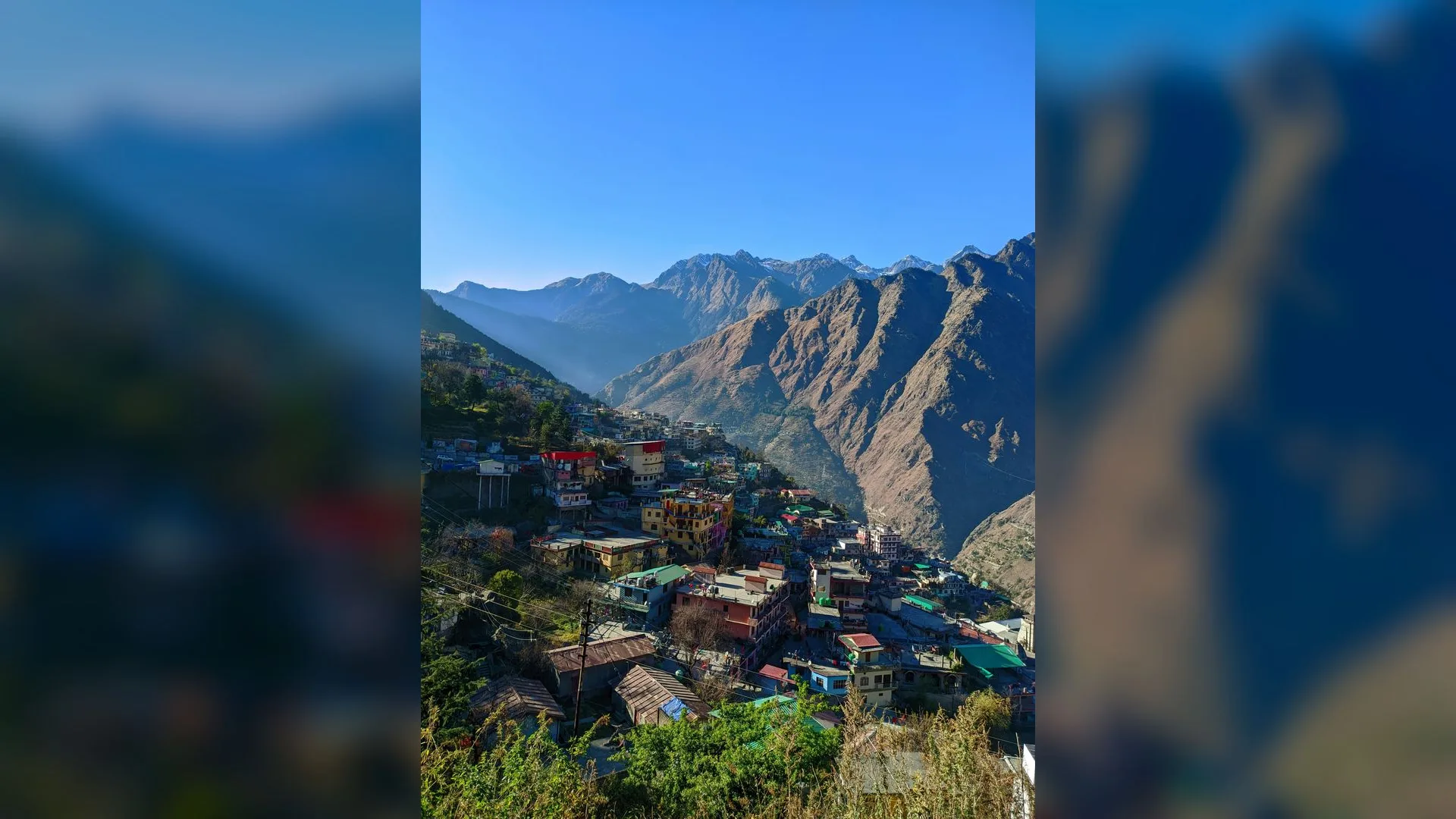 Dağ yamacına kurulmuş renkli binalı kasaba, arkada sarp kayalık dağlar ve açık mavi gökyüzü