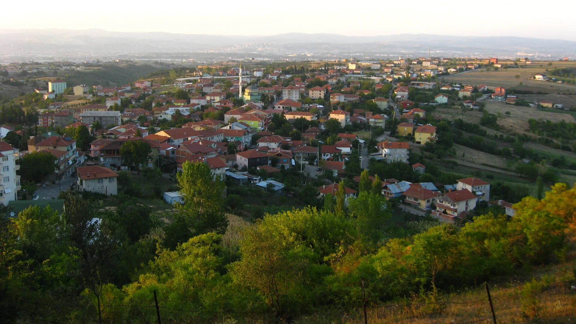 Kırmızı çatılı evler ve minareli yeşil bir yerleşimin tepeden görünümü, arkada sisli dağlar
