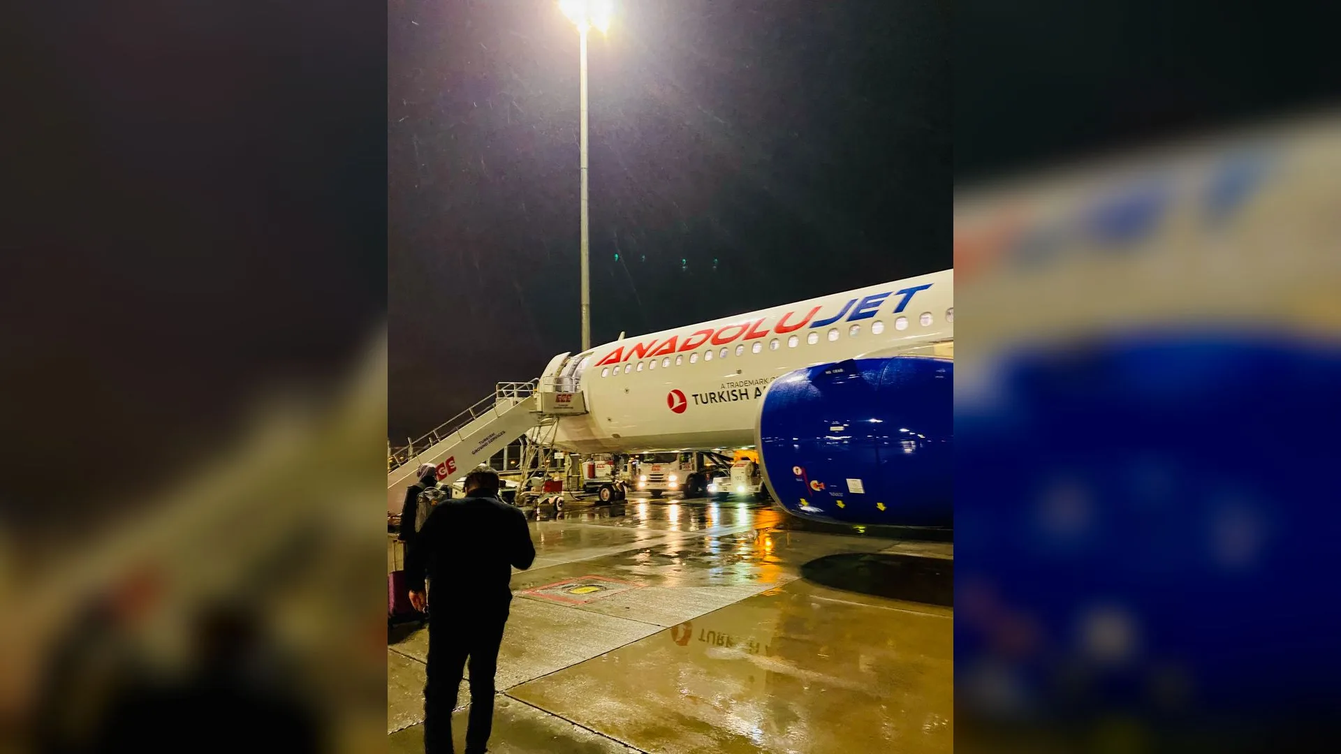 Yağmurlu bir gecede apronda duran AnadoluJet uçağı, merdiveni ve önünde yürüyen bir adam