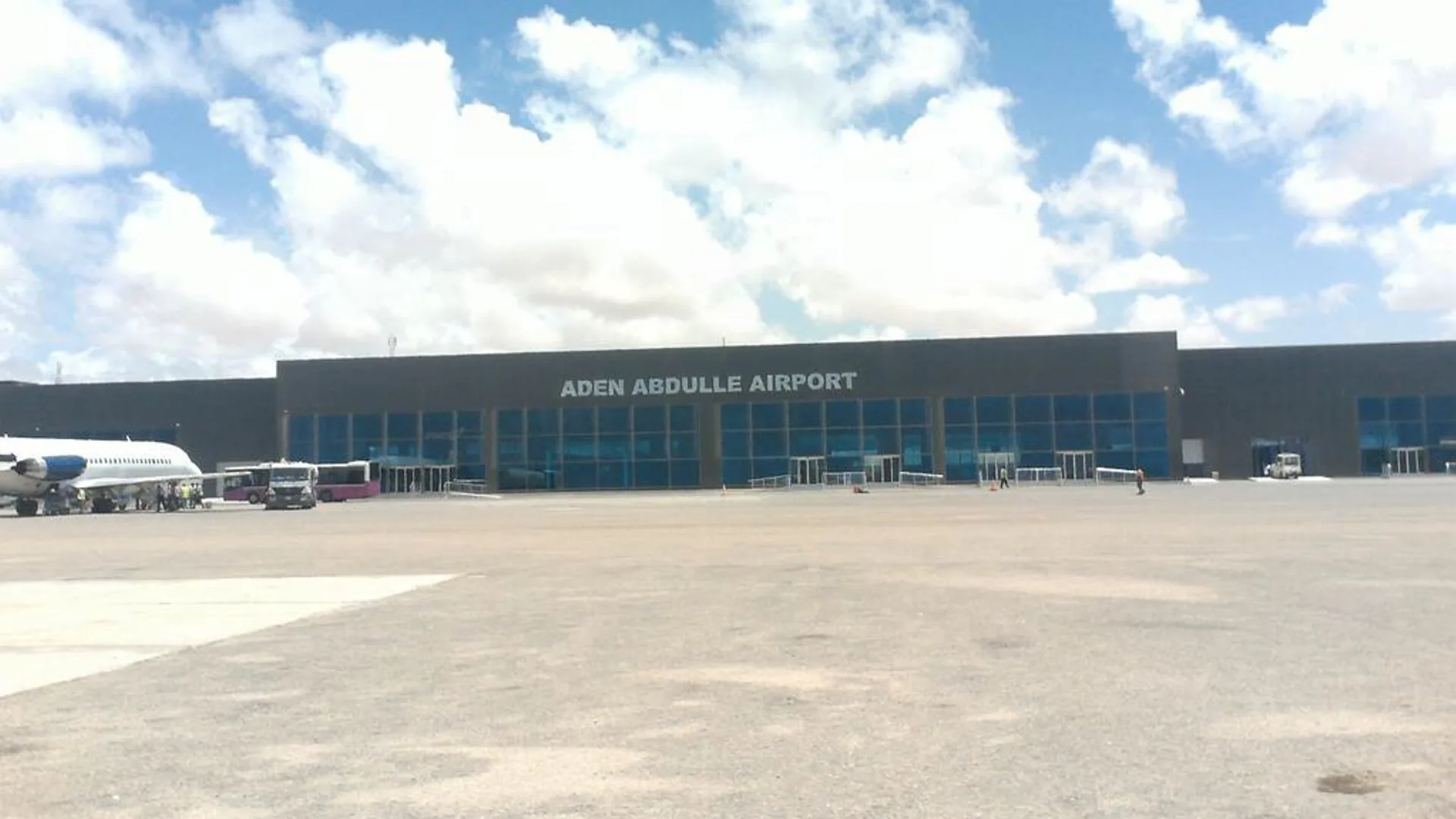 Bulutlu gökyüzü altında, cephesinde 'Aden Abdulle Airport' yazan terminal binası ve apronda bekleyen bir yolcu uçağı