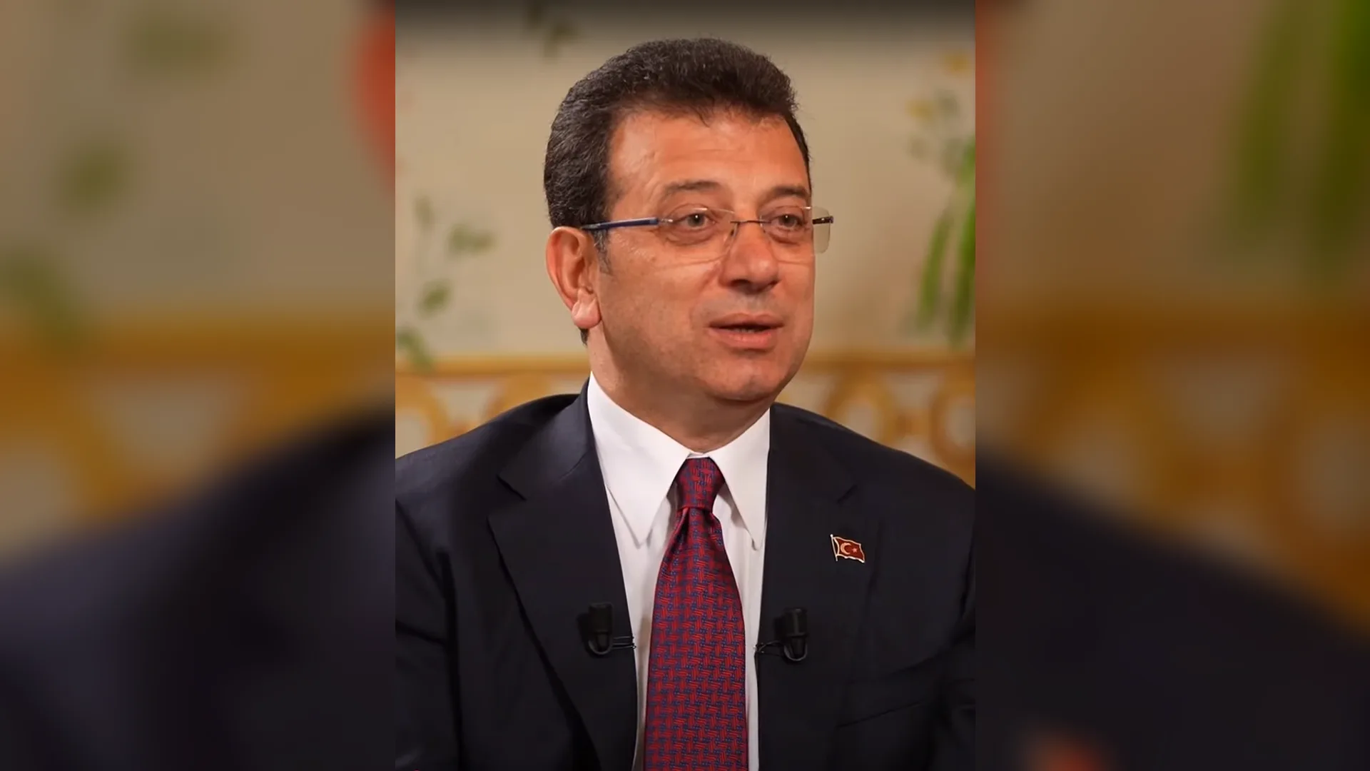 Gözlüklü, koyu takım elbiseli ve bordo kravatlı bir adam konuşurken