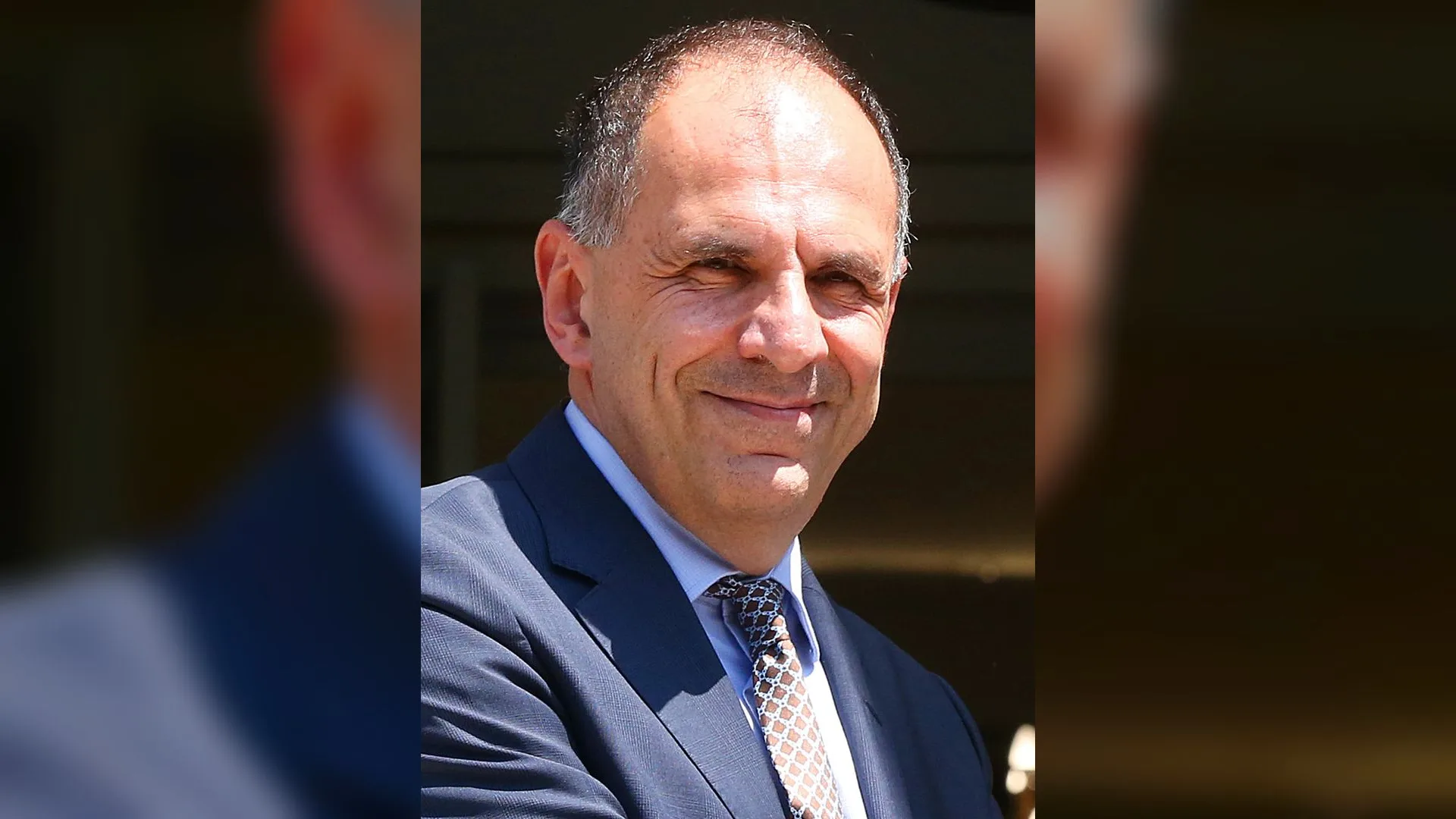 Lacivert takım elbiseli, desenli kravatlı, kısa saçlı gülümseyen orta yaşlı bir adam