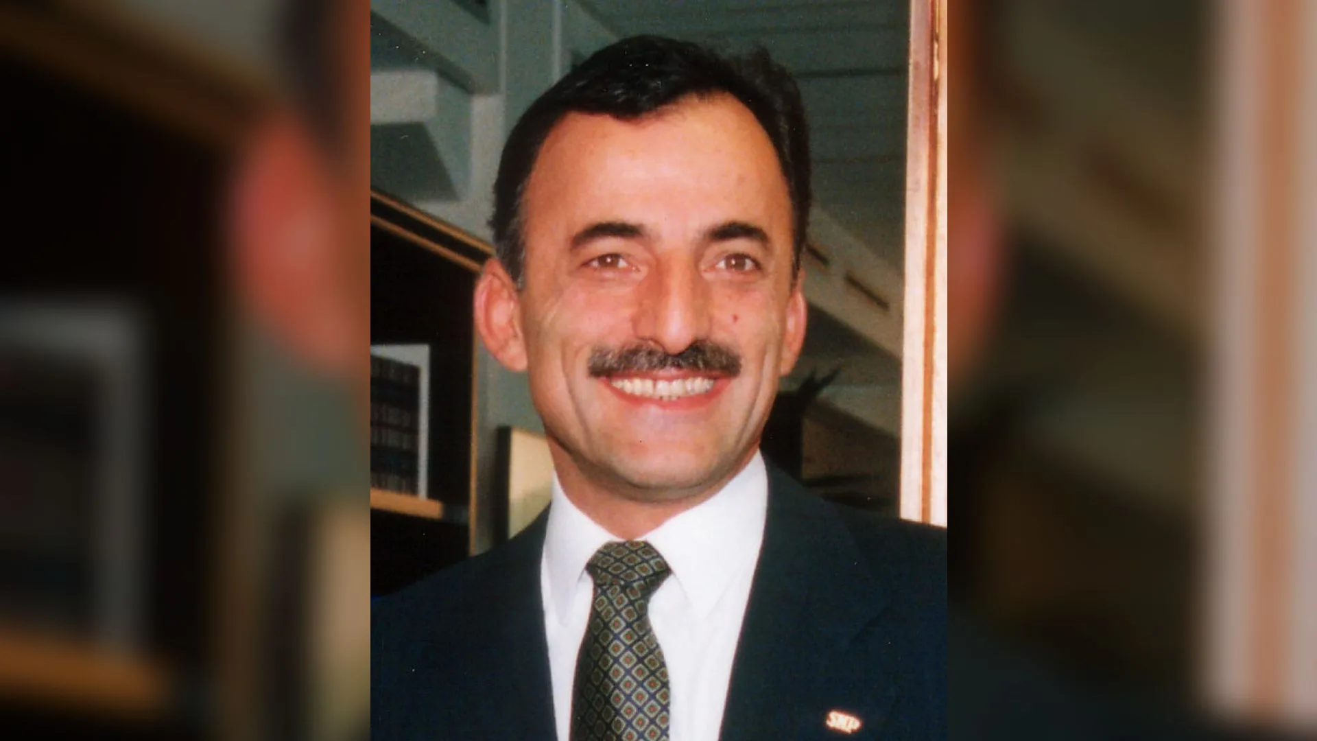 Koyu takım elbiseli, desenli kravatlı, bıyıklı ve gülümseyen bir adamın portresi