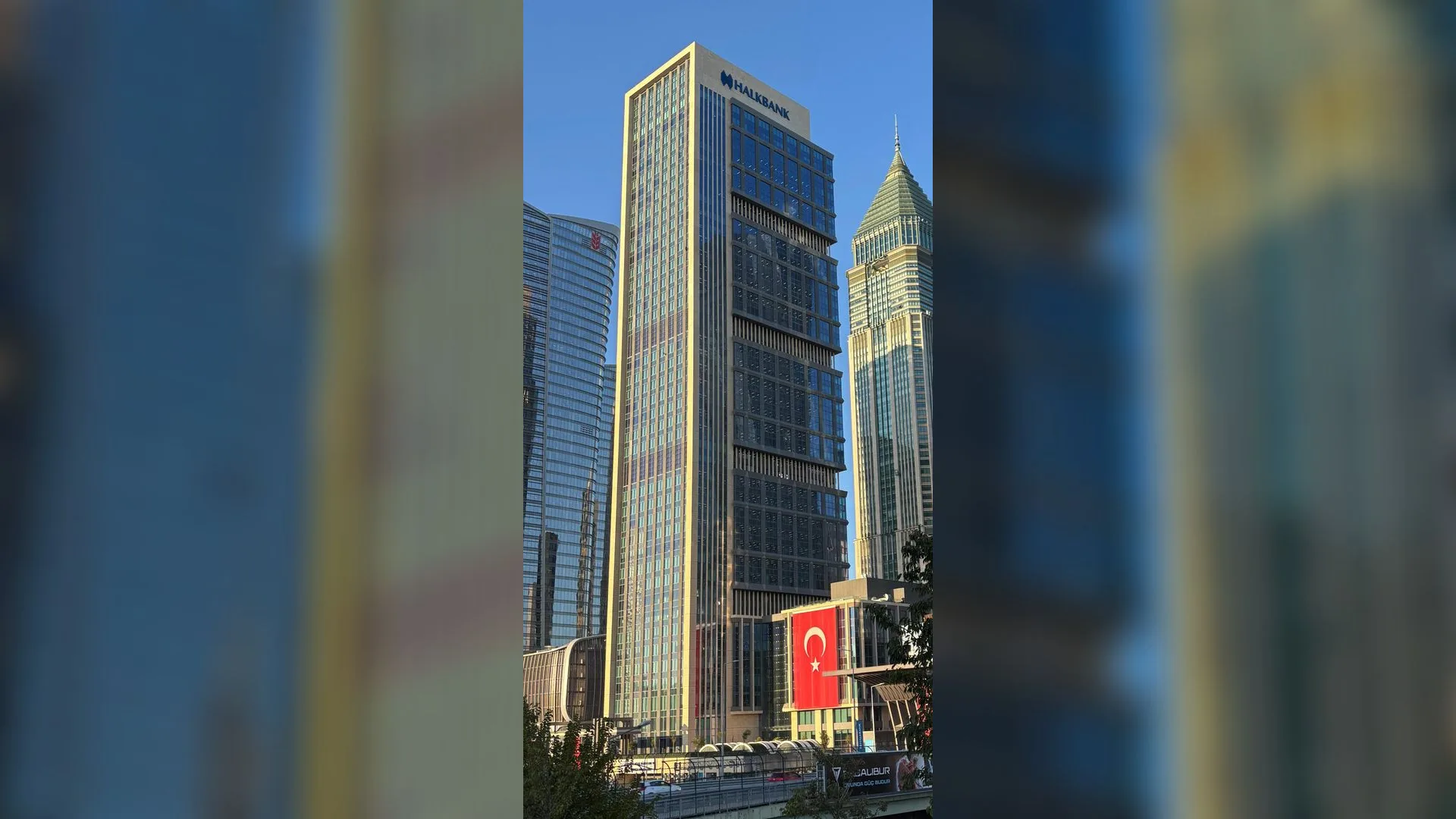 Mavi gökyüzü altında Halkbank logolu cam gökdelen ve önünde asılı Türk bayrağı