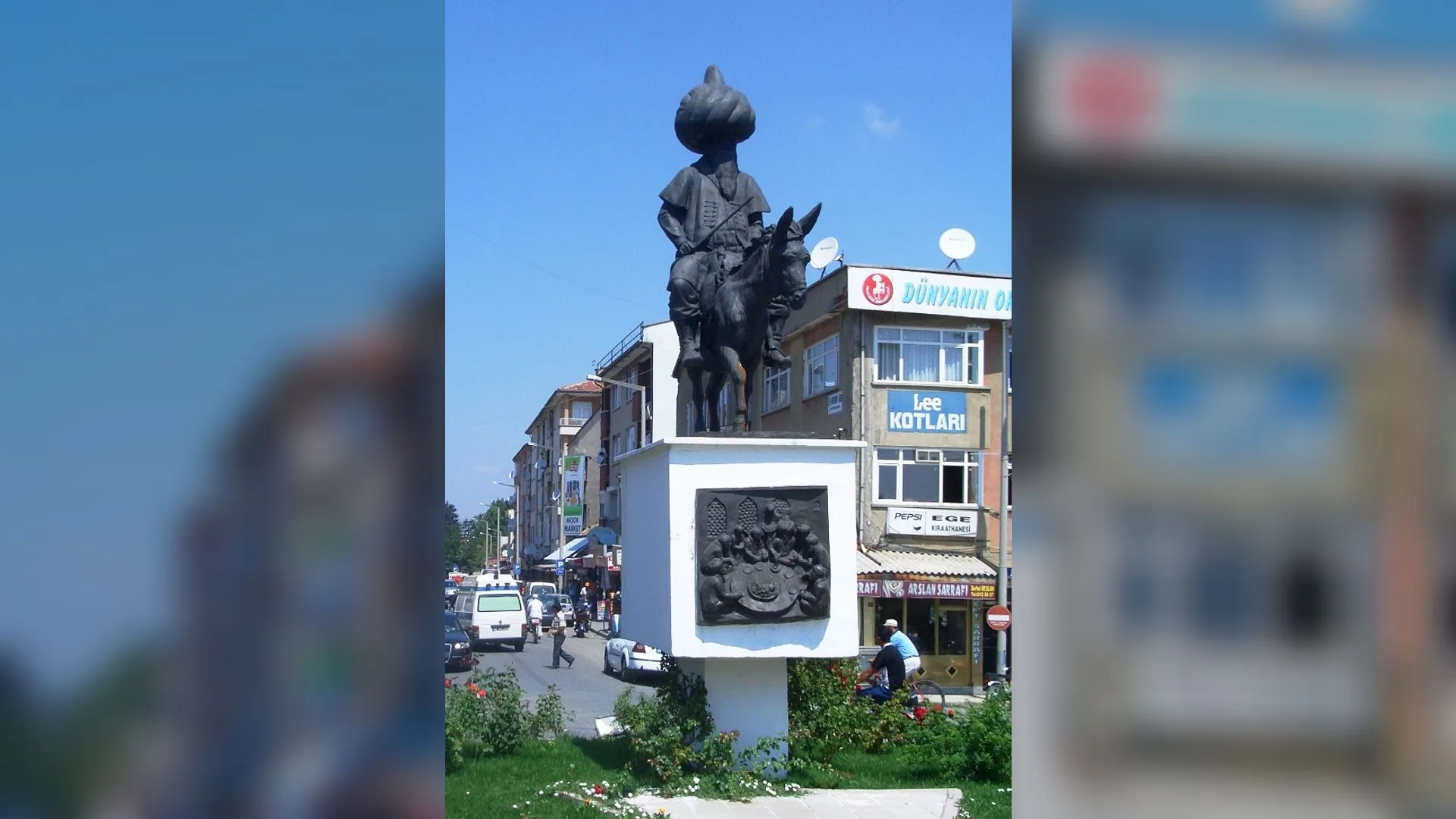 Şehir meydanında eşeğe ters binmiş kavuklu bir adam heykeli, arkada dükkanlar ve binalar