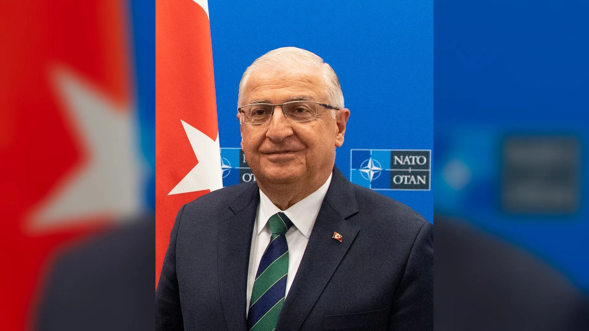 Türk bayrağı ve NATO logosu önünde gözlüklü, takım elbiseli beyaz saçlı bir adam