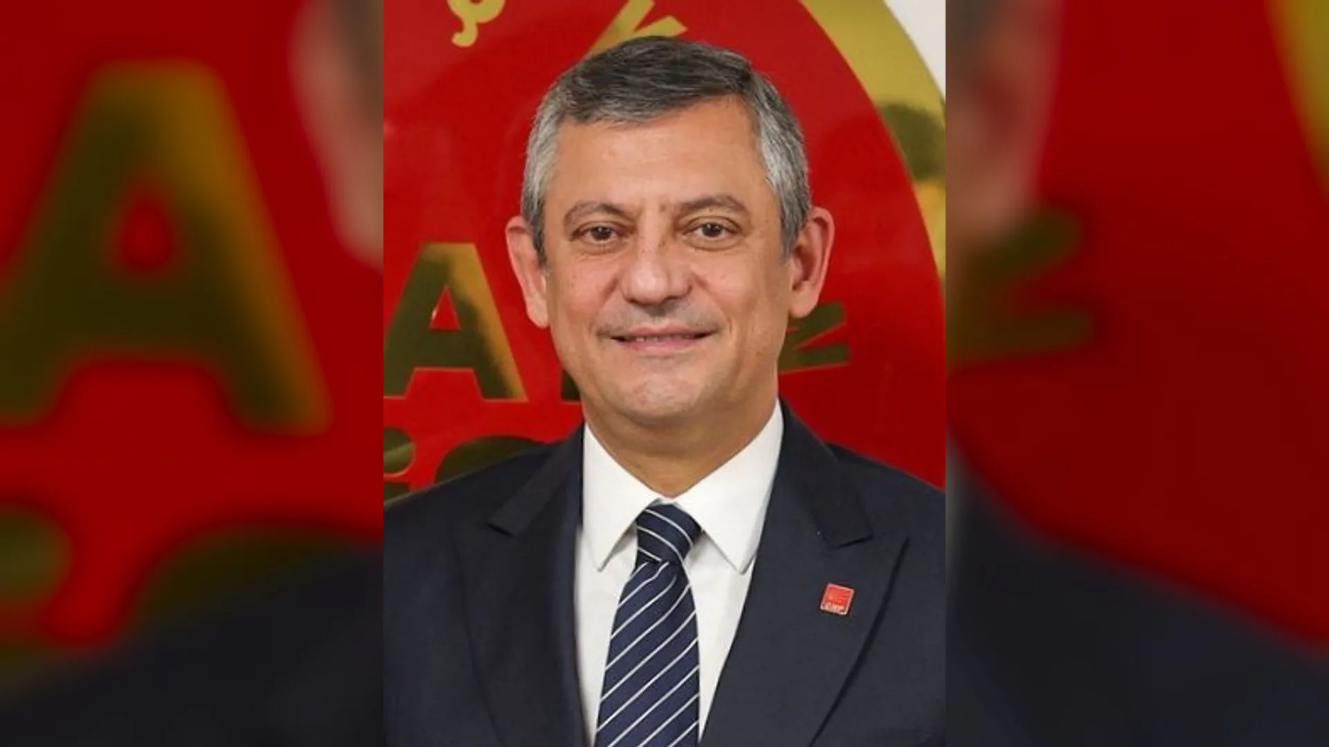 Kırmızı arka plan önünde lacivert takım elbiseli, çizgili kravatlı, gülümseyen gri saçlı adam