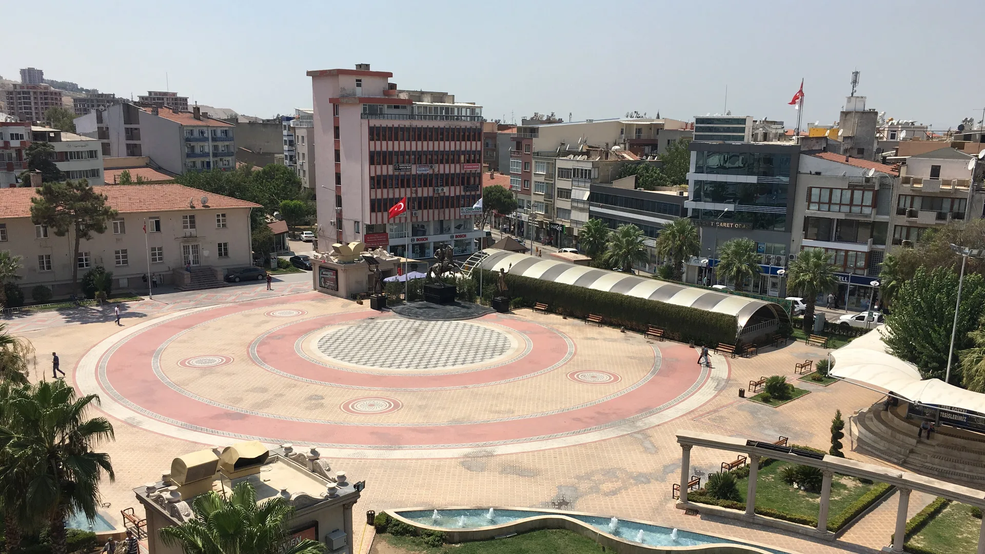 Palmiyeler ve binalarla çevrili, desenli zemini olan geniş bir kent meydanı ile süs havuzu