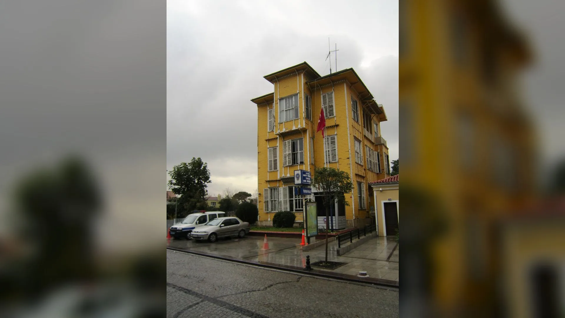Yağmurlu havada önünde araçlar park etmiş, Türk bayraklı sarı ahşap karakol binası