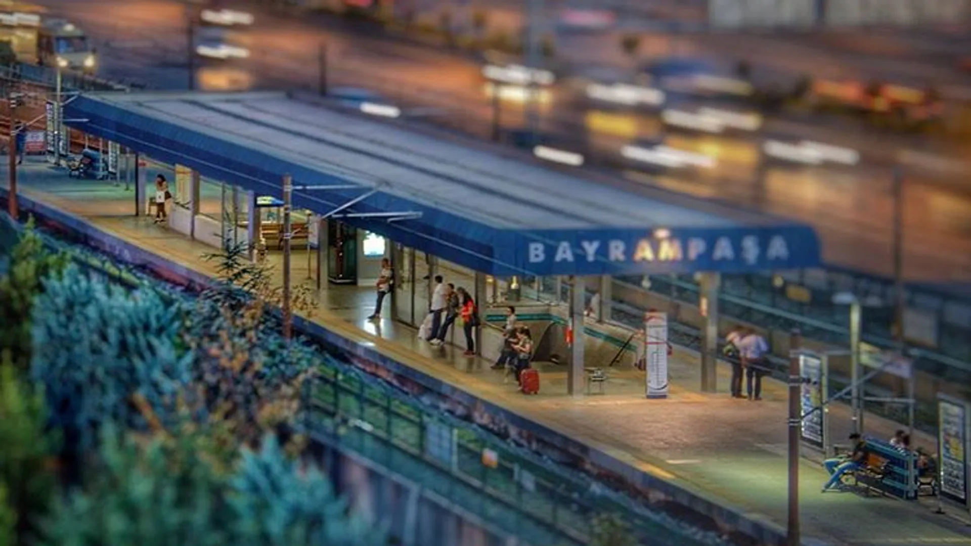 Mavi çatılı Bayrampaşa metro istasyonu peronunda bekleyen yolcular, arkada akşam trafiği