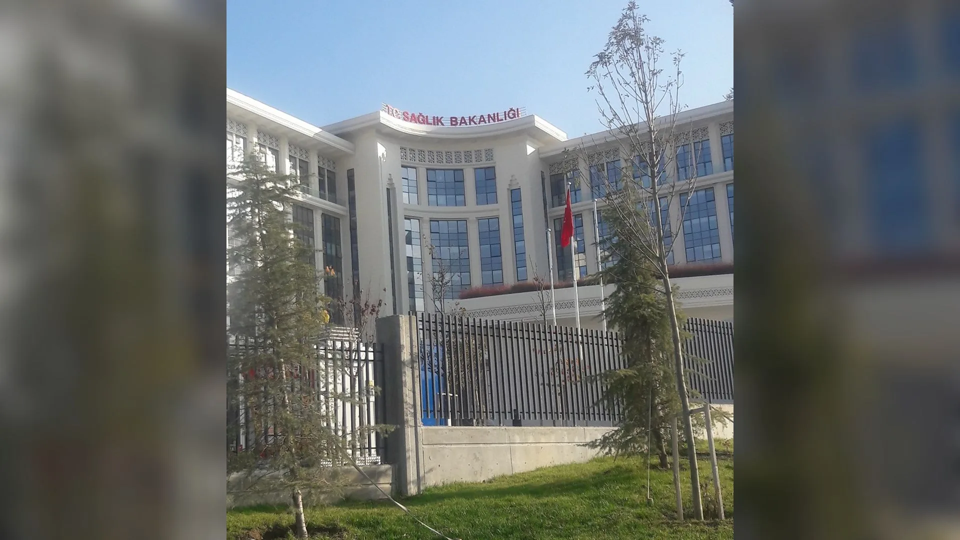Çatısında Sağlık Bakanlığı yazan beyaz bina, önünde Türk bayrağı, demir çit ve çimenlik