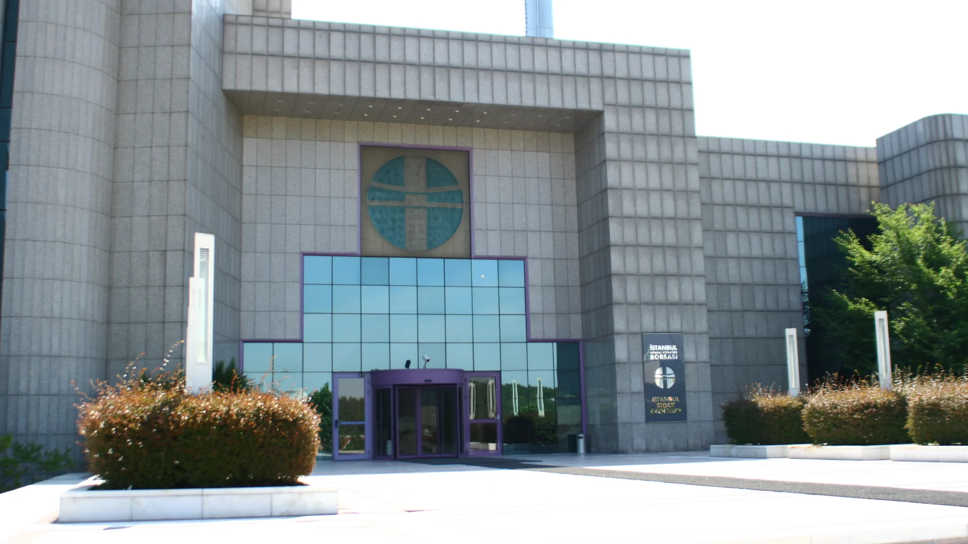 Taş kaplı, cam girişli modern bir binanın önü ve çalılar