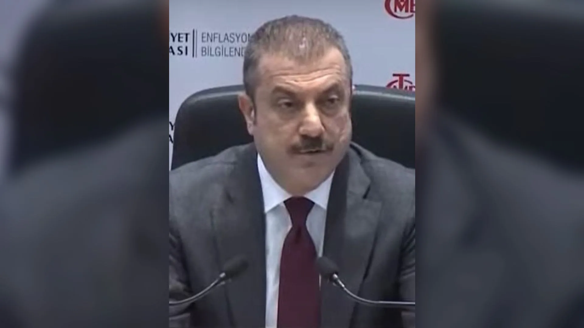Bıyıklı, takım elbiseli ve bordo kravatlı bir adam mikrofonların önünde konuşuyor