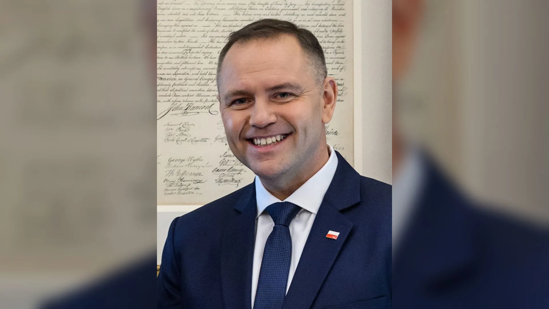 Lacivert takım elbiseli, gülümseyen bir adam, arkasında el yazısı bir belge çerçevesi.
