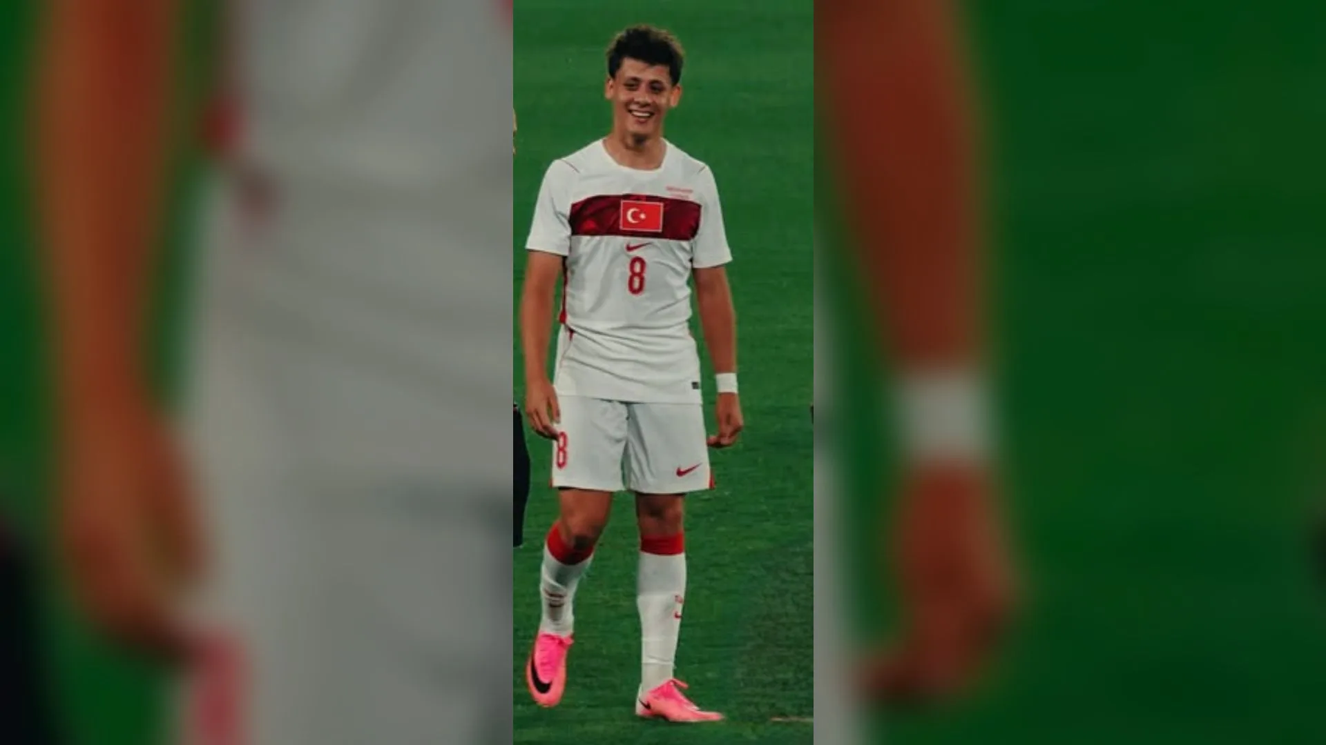 Beyaz 8 numaralı milli forma giymiş, pembe kramponlu gülümseyen genç futbolcu sahada yürüyor