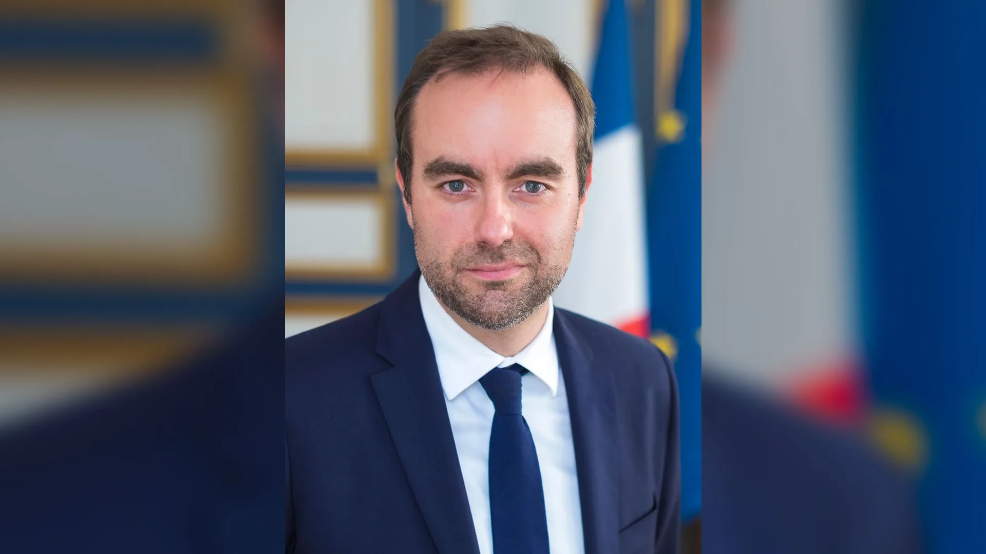 Fransız ve AB bayrakları önünde lacivert takım elbiseli sakallı adam