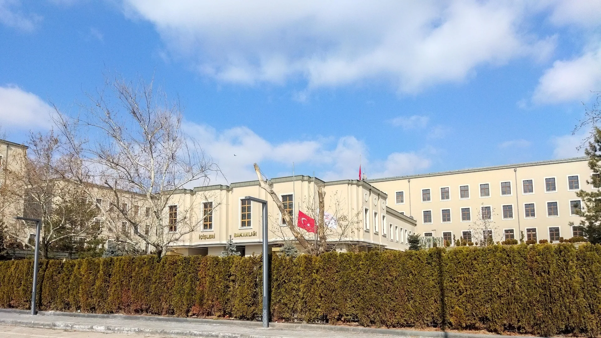 Çit arkasında Türk bayrağı dalgalanan, krem renkli büyük bir resmi bina ve önünde boş bir cadde