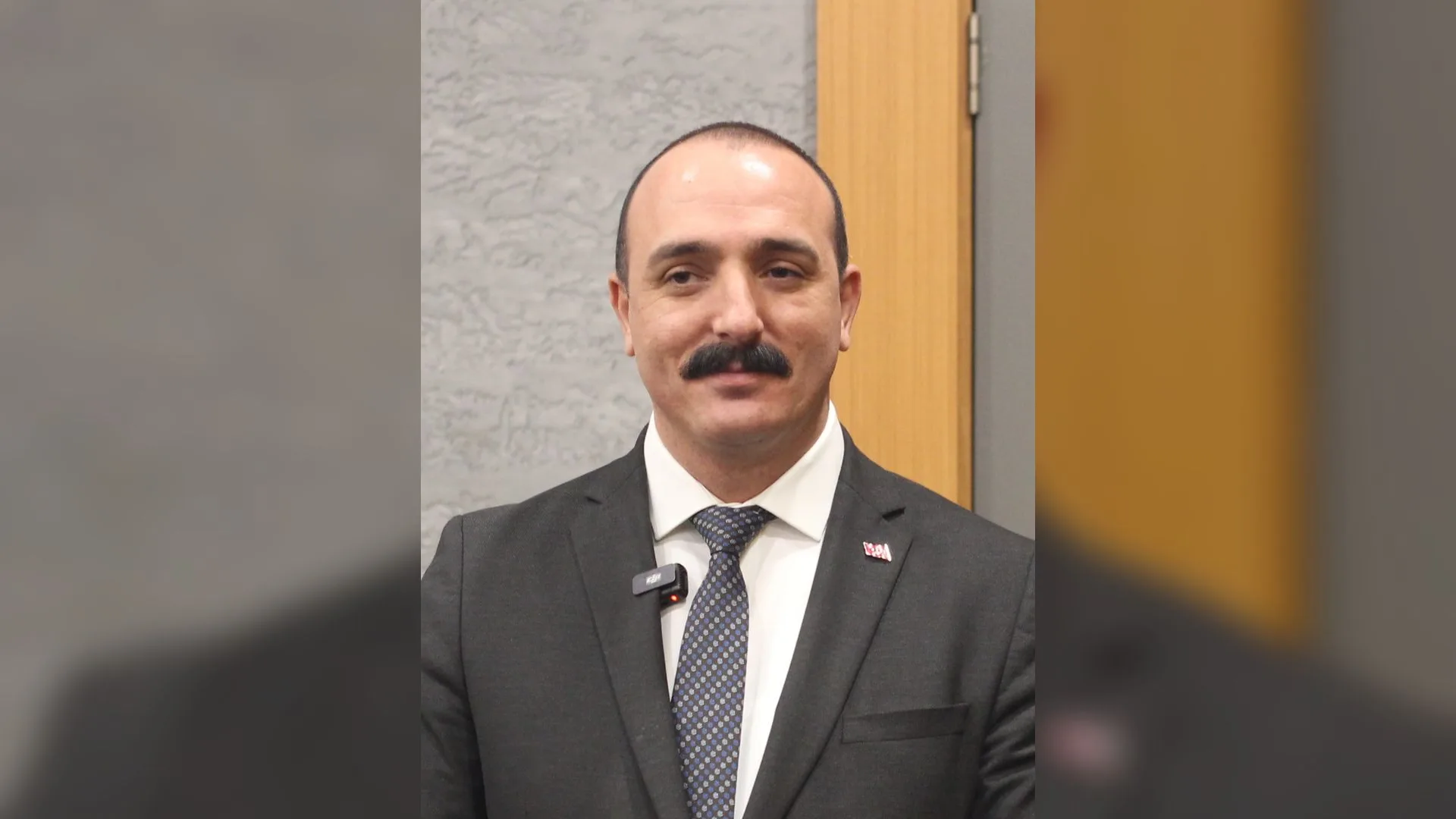 Gri takım elbiseli, kravatlı, bıyıklı bir adam gülümseyerek poz veriyor.