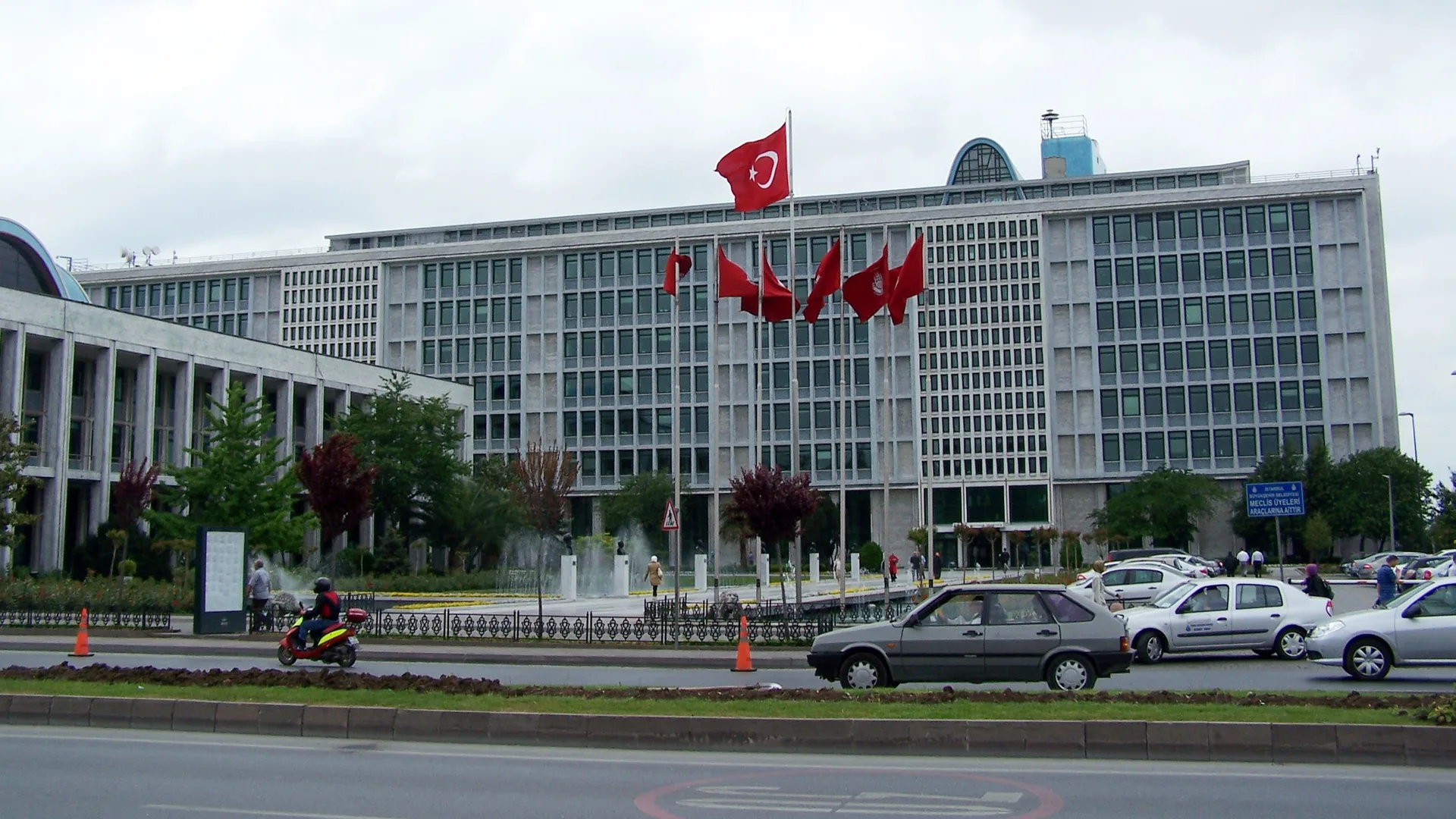 Önünde Türk bayrakları dalgalanan çok katlı beyaz bir bina ve önündeki yoldan geçen arabalar