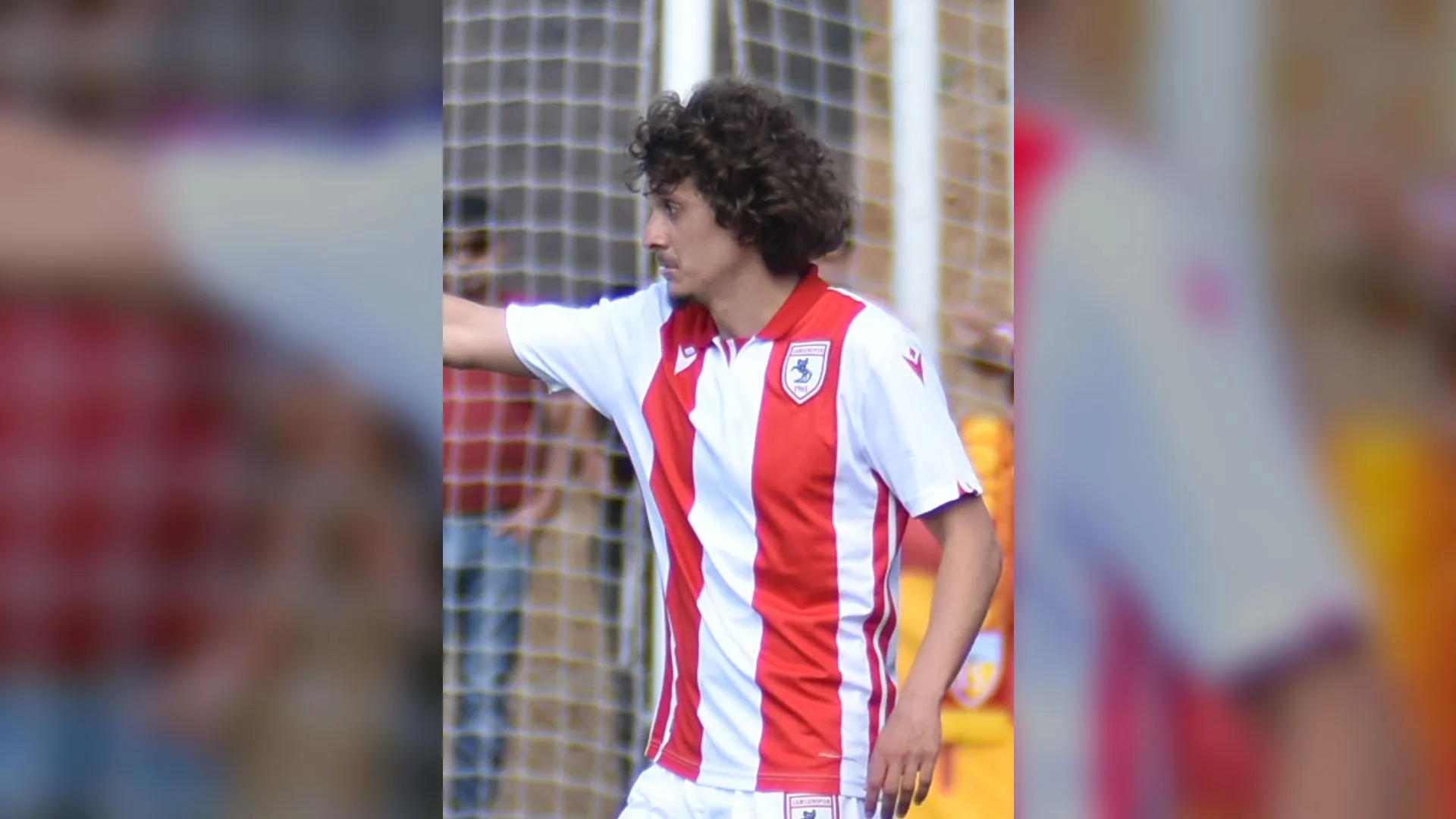 Kıvırcık saçlı, kırmızı-beyaz çizgili formalı bir futbolcu kale ağları önünde kolunu uzatıyor
