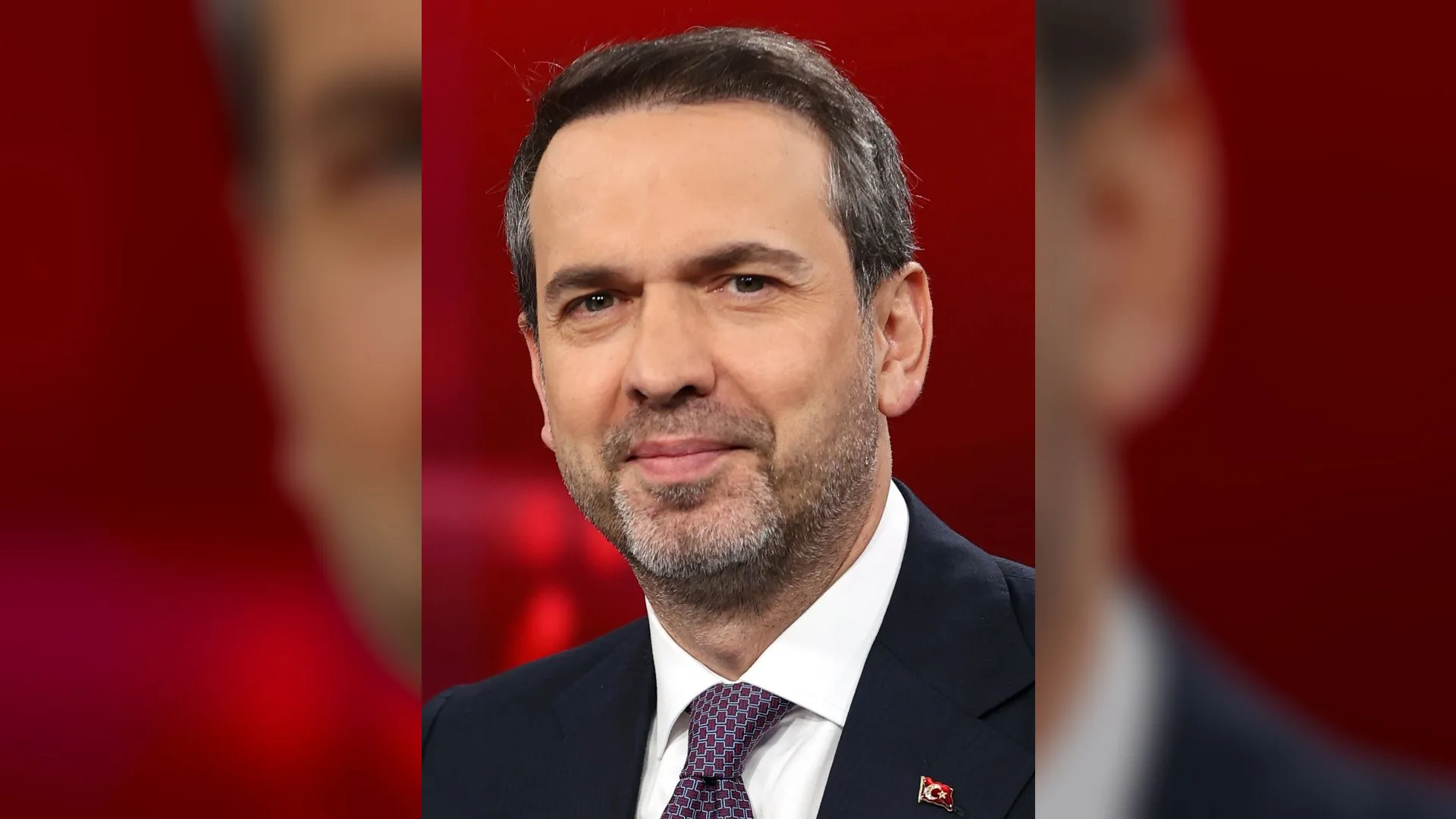 Kırmızı arka plan önünde lacivert takım elbiseli, mor kravatlı, kırçıl sakallı gülümseyen bir adam