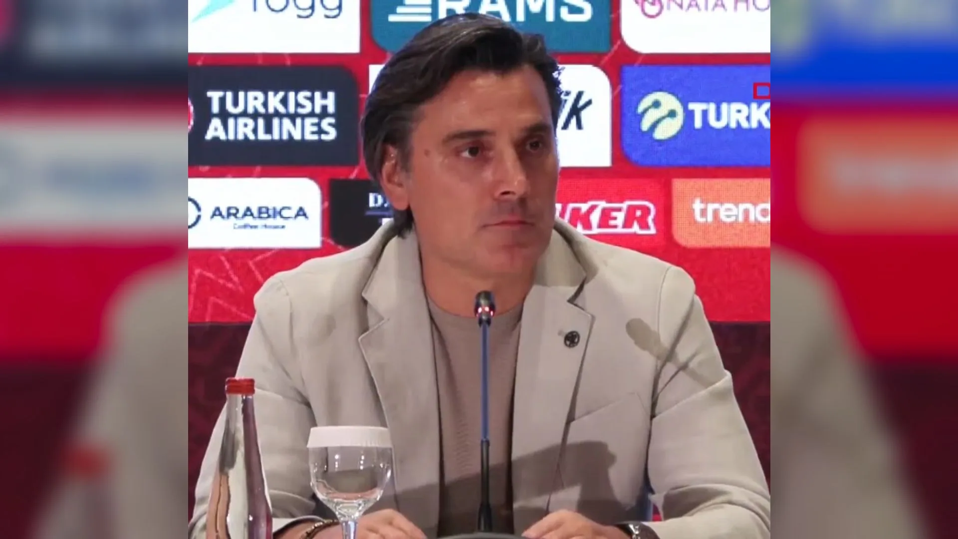 Sponsor logolu panonun önünde mikrofon başında oturan, açık renk ceketli koyu saçlı bir adam