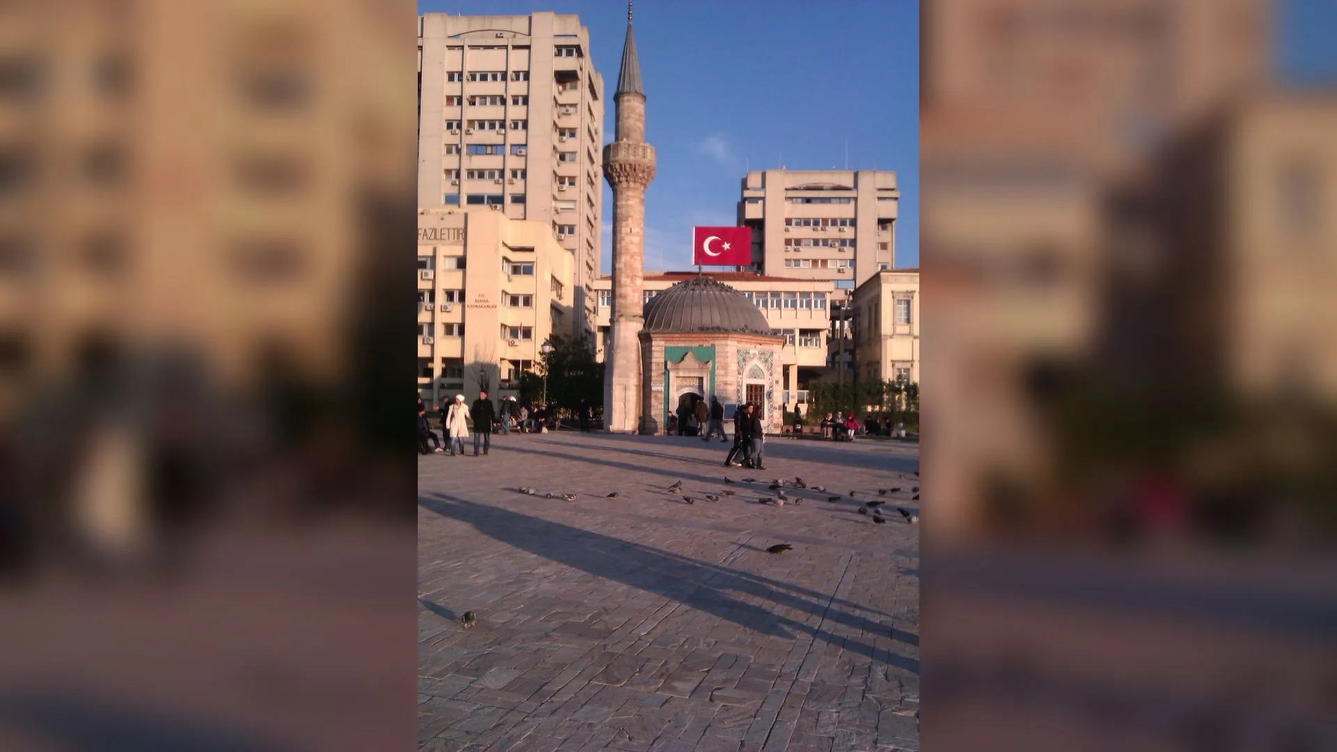 Konak Meydanı'nda minareli küçük cami, Türk bayraklı binalar ve güvercinler