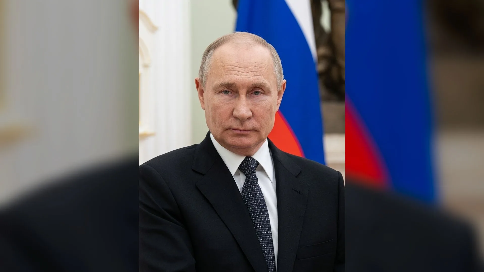 Rus bayrağı önünde koyu takım elbise ve lacivert kravat giymiş kır saçlı bir adam