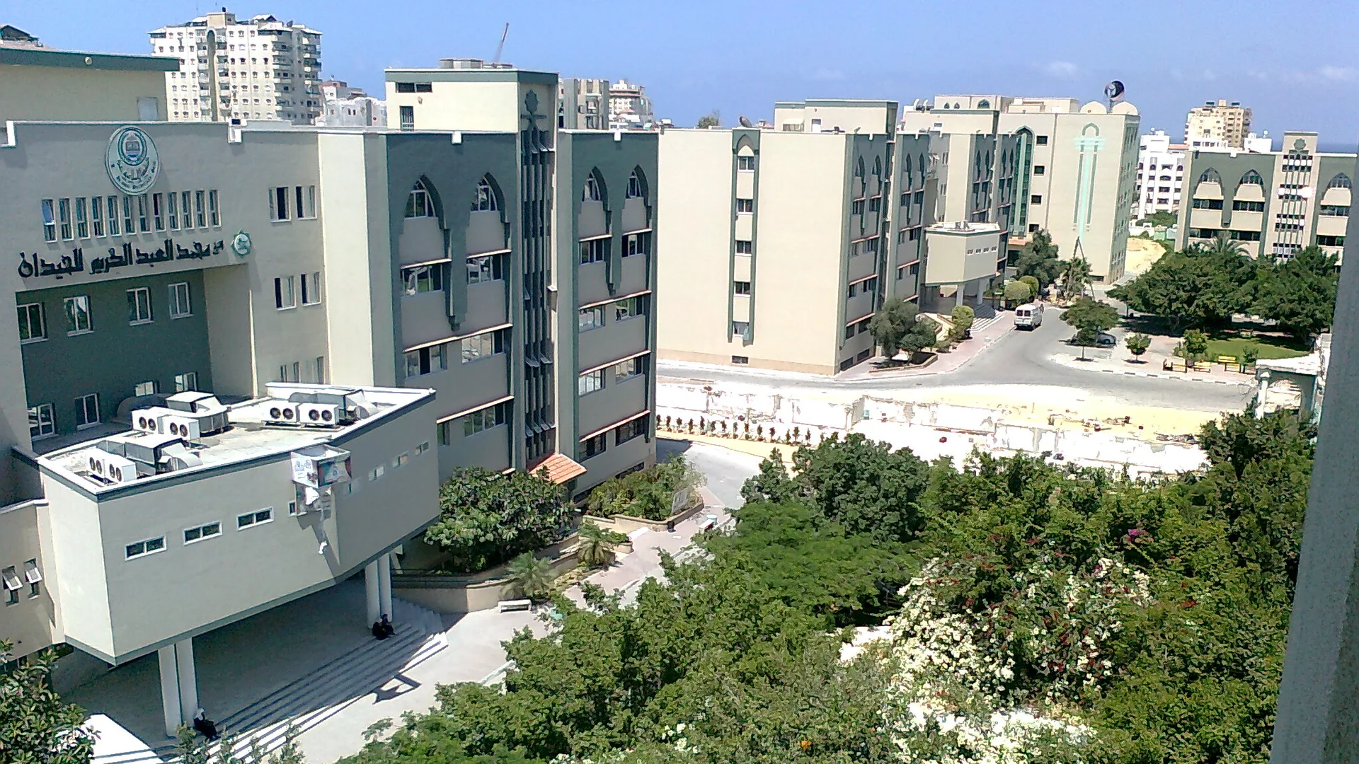 Ağaçlarla çevrili, kemerli pencereli bej renkli çok katlı kampüs binaları ve aralarındaki yol