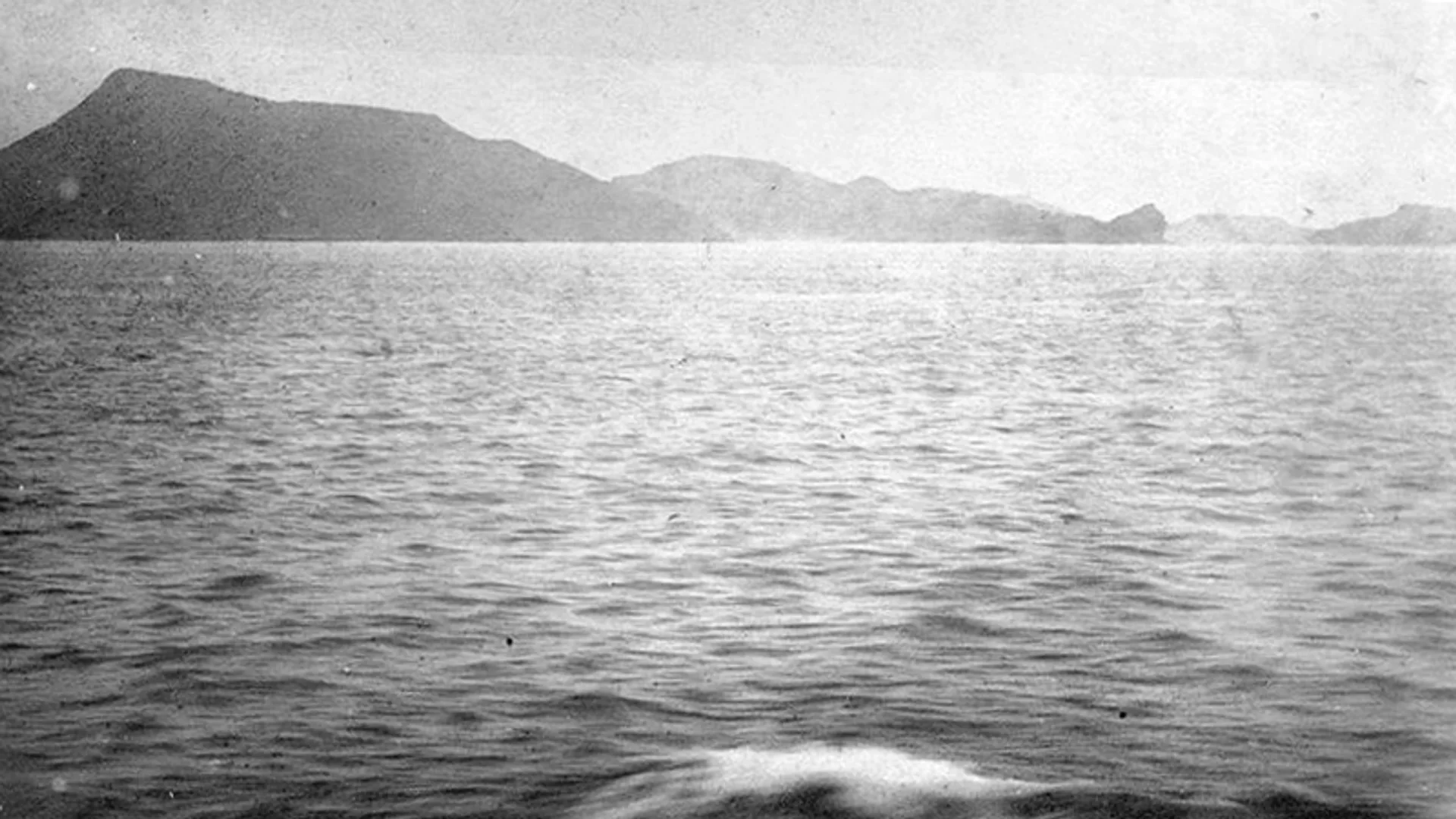 Siyah beyaz eski bir karede dalgalı deniz ve ufukta dağlık kıyı şeridi