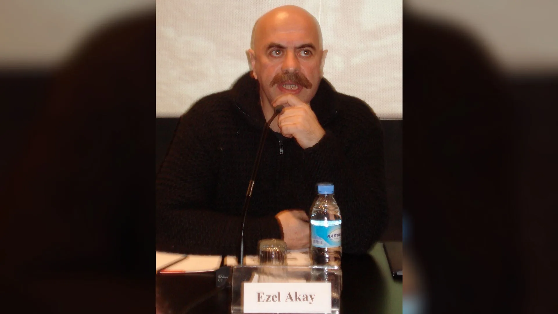Siyah kazaklı, bıyıklı kel bir adam masada mikrofon önünde oturuyor; önünde "Ezel Akay" yazılı isimlik var.