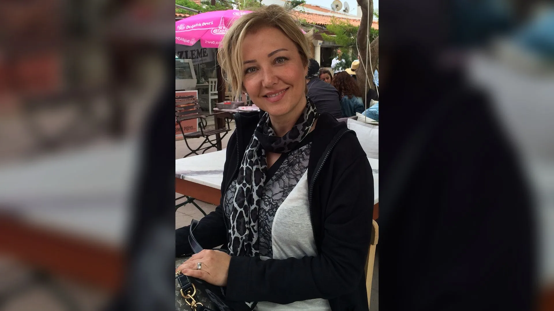 Desenli fular ve siyah hırka giymiş sarışın bir kadın, açık hava kafesinde gülümsüyor.