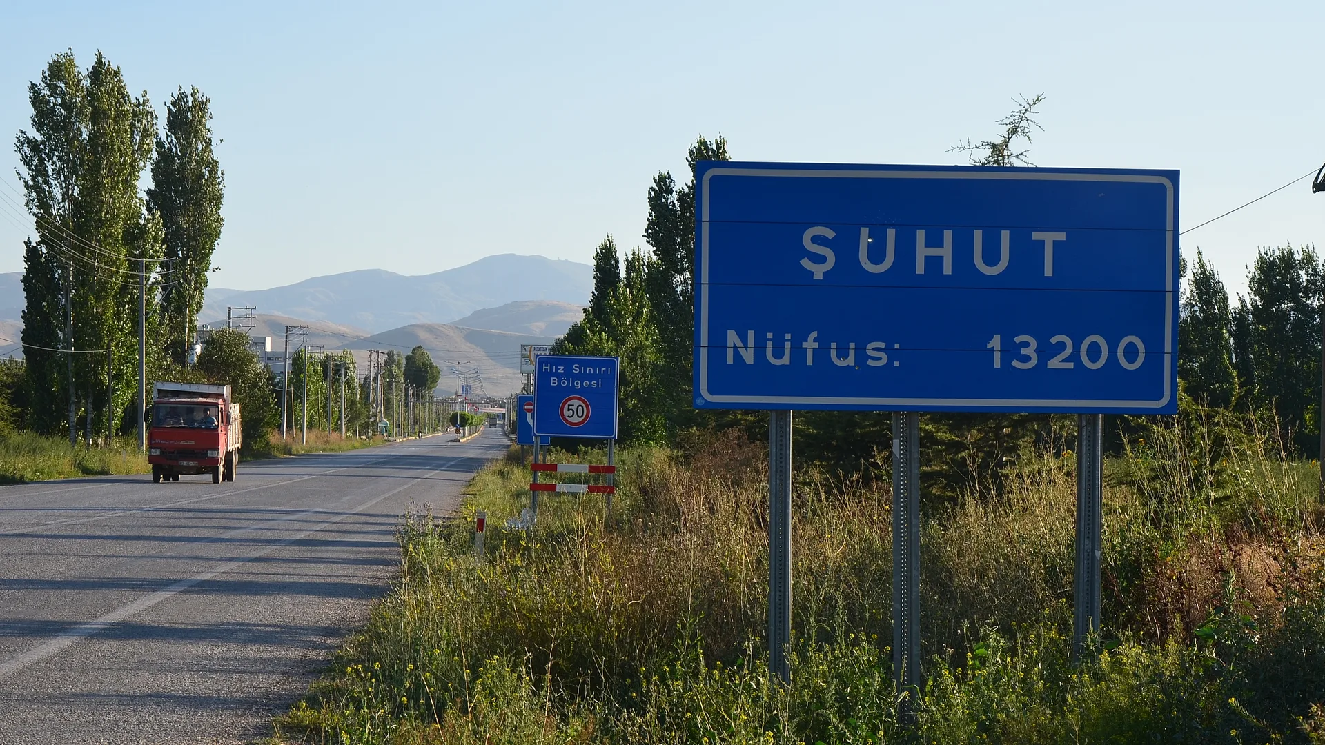 Yol kenarında mavi Şuhut nüfus tabelası, yolda kırmızı bir kamyon ve arkada dağlar