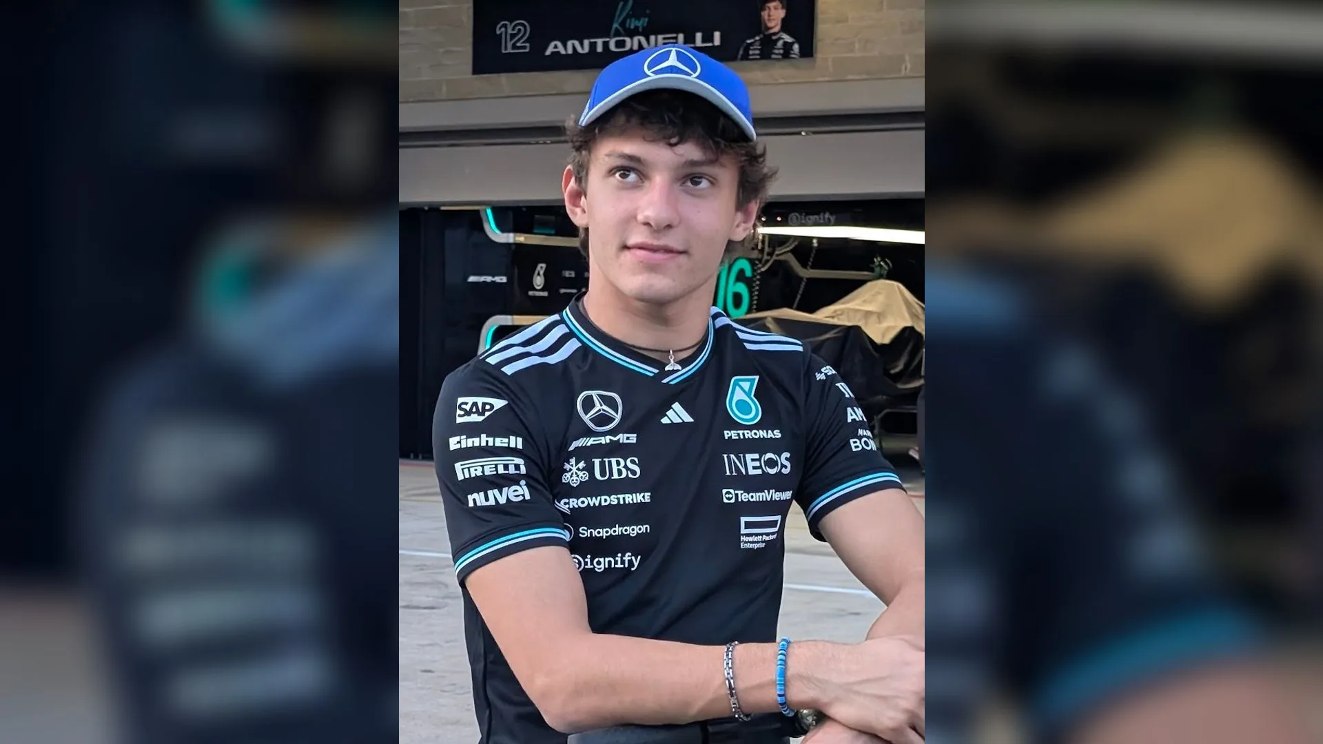 Mavi şapkalı, Mercedes takım tişörtlü genç bir pilot, kolları kavuşturmuş halde pit garajı önünde duruyor.
