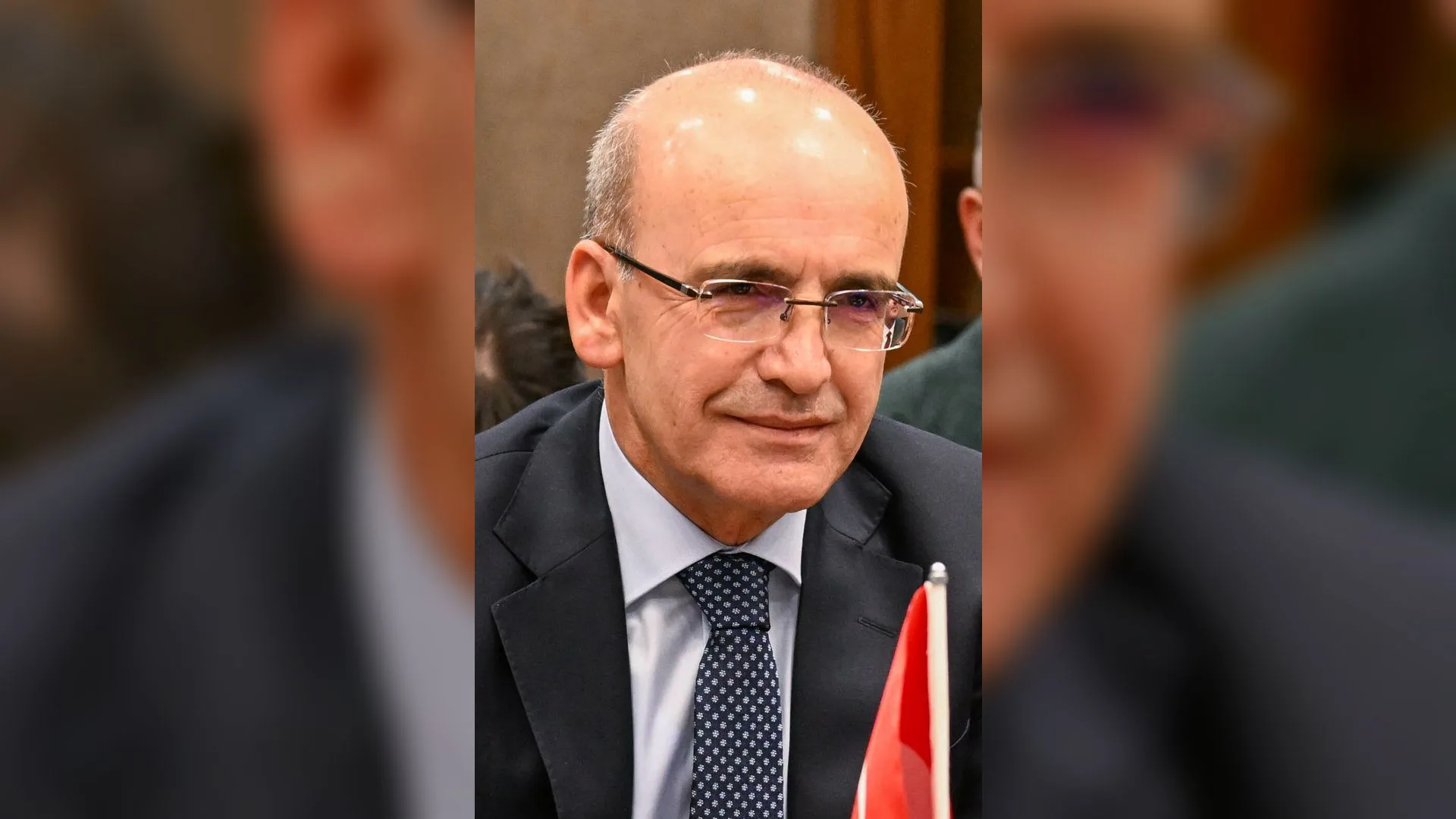 Gözlüklü, takım elbiseli ve kravatlı kel bir adam gülümsüyor; önünde küçük bir Türk bayrağı duruyor.