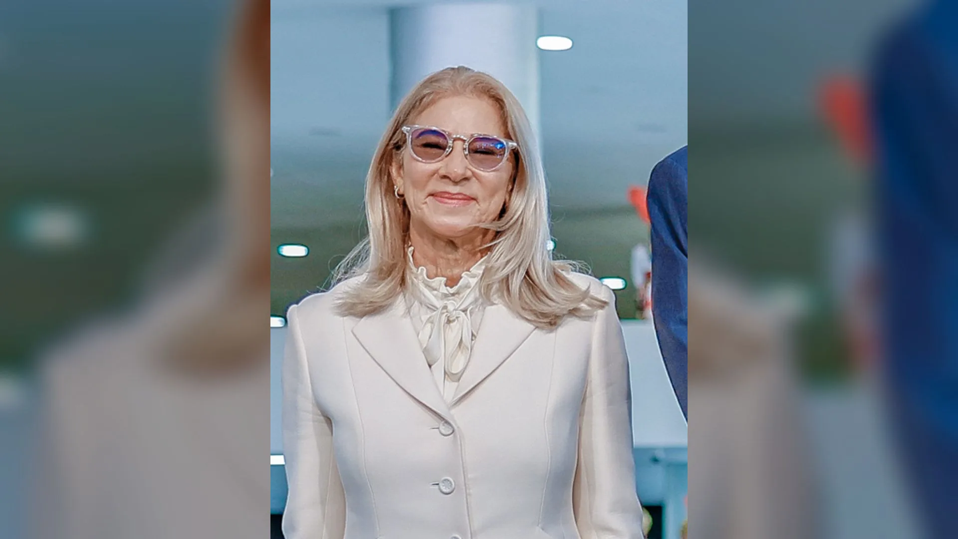 Beyaz ceketli, renkli camlı gözlüklü, uzun sarı saçlı gülümseyen bir kadın
