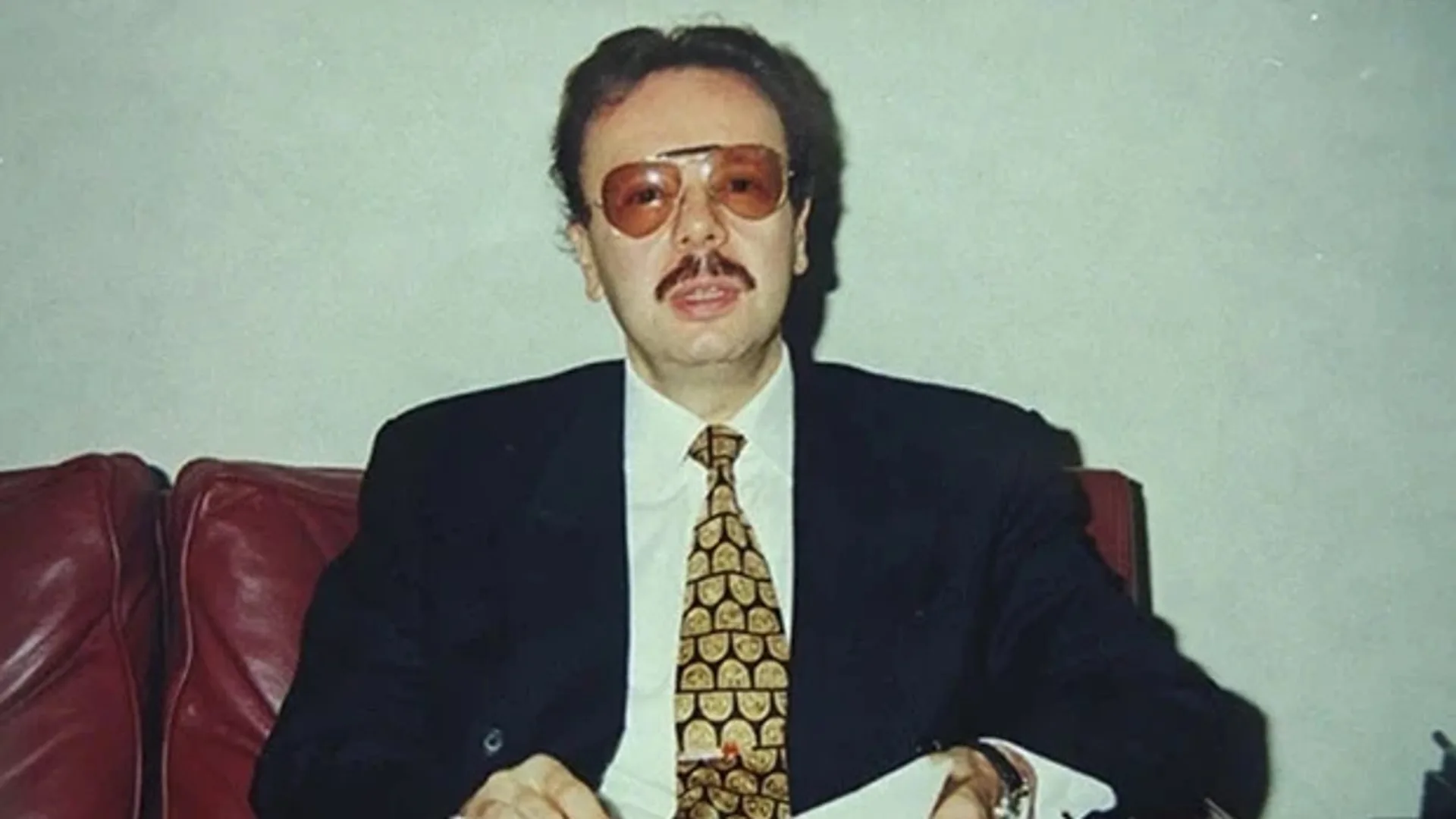 Renkli gözlüklü, bıyıklı, takım elbiseli ve desenli kravatlı bir adam kırmızı deri koltukta oturuyor