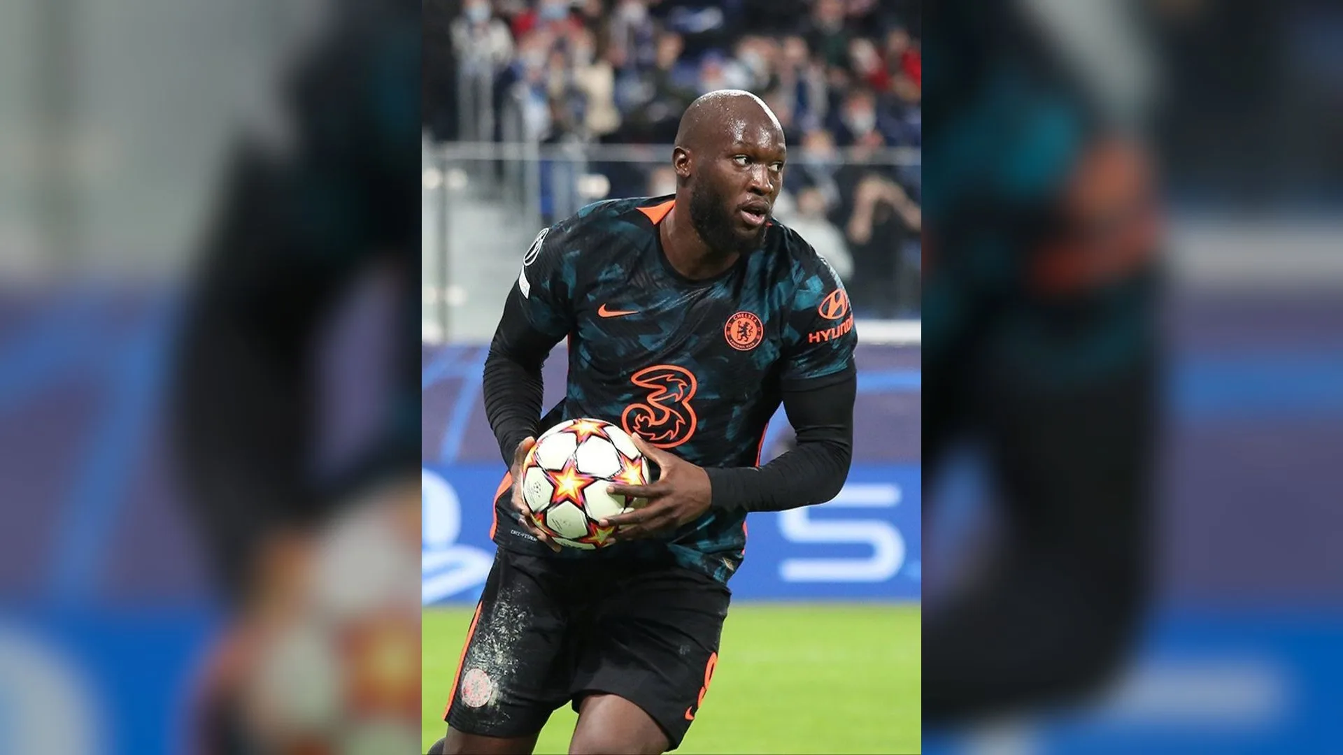 Koyu renkli forma giymiş bir futbolcu, elinde topla sahada yürüyor