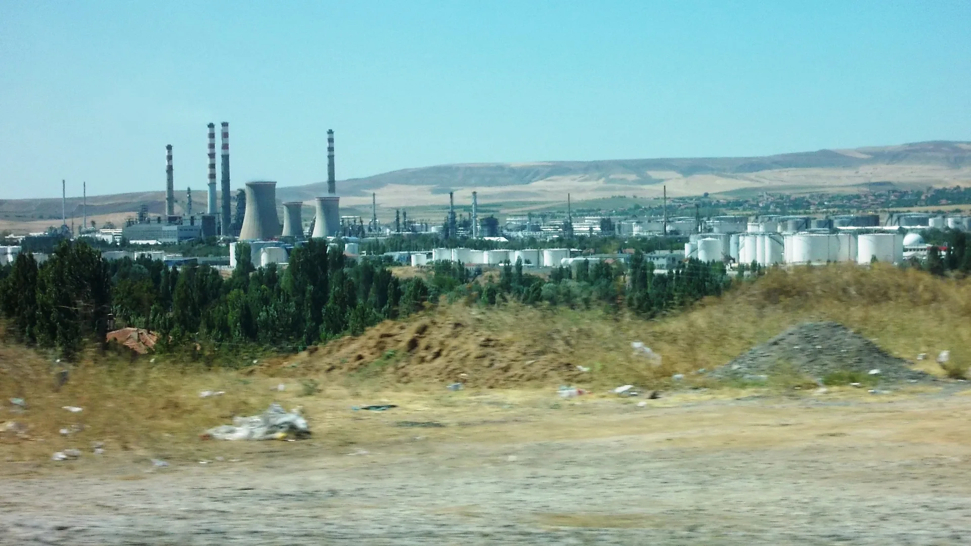 Uzakta bacaları ve beyaz depolama tanklarıyla bir rafineri, önde kuru otlu boş arazi