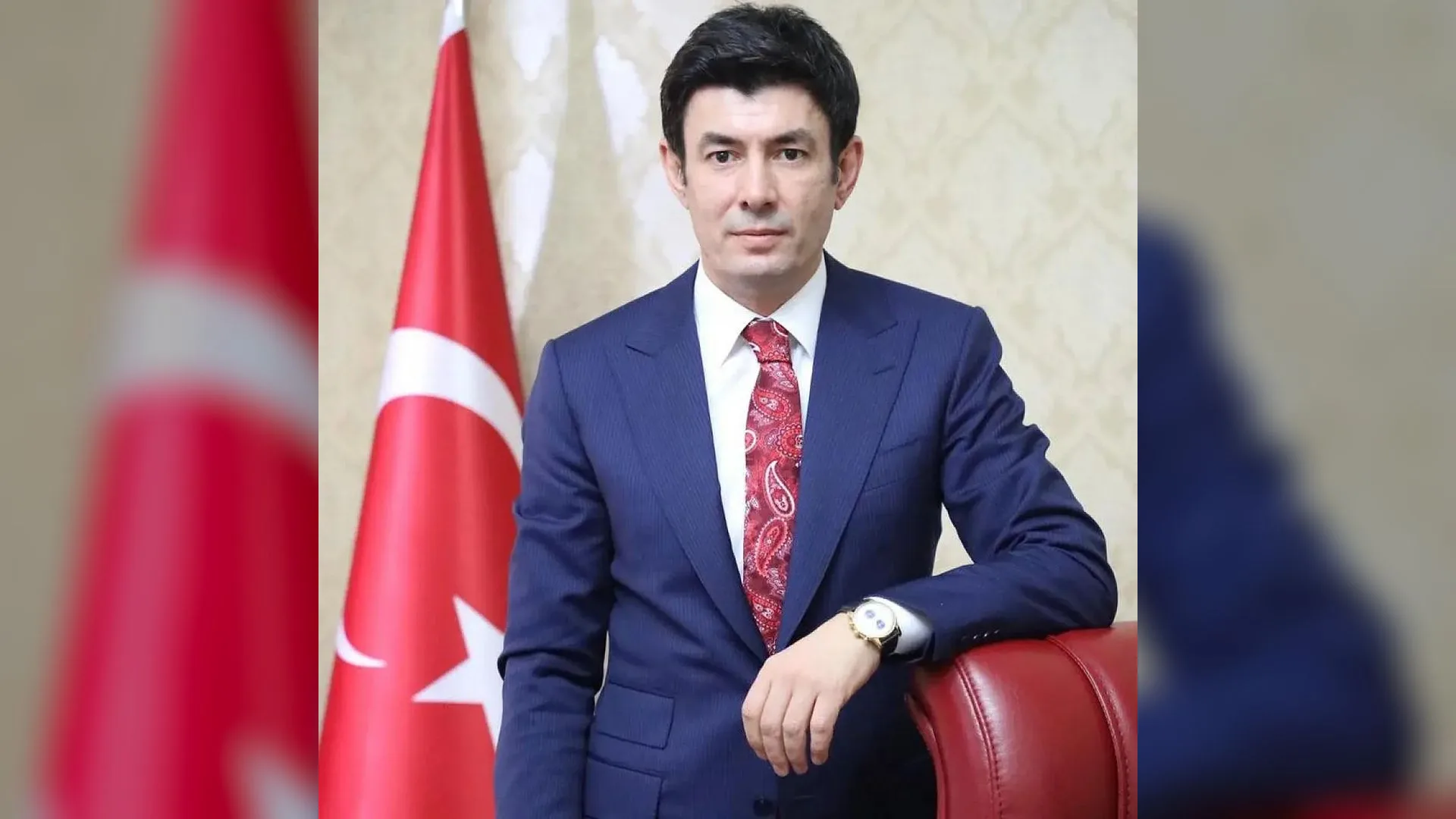 Lacivert takım elbiseli, kırmızı kravatlı bir adam Türk bayrağı önünde deri koltuğa yaslanmış.