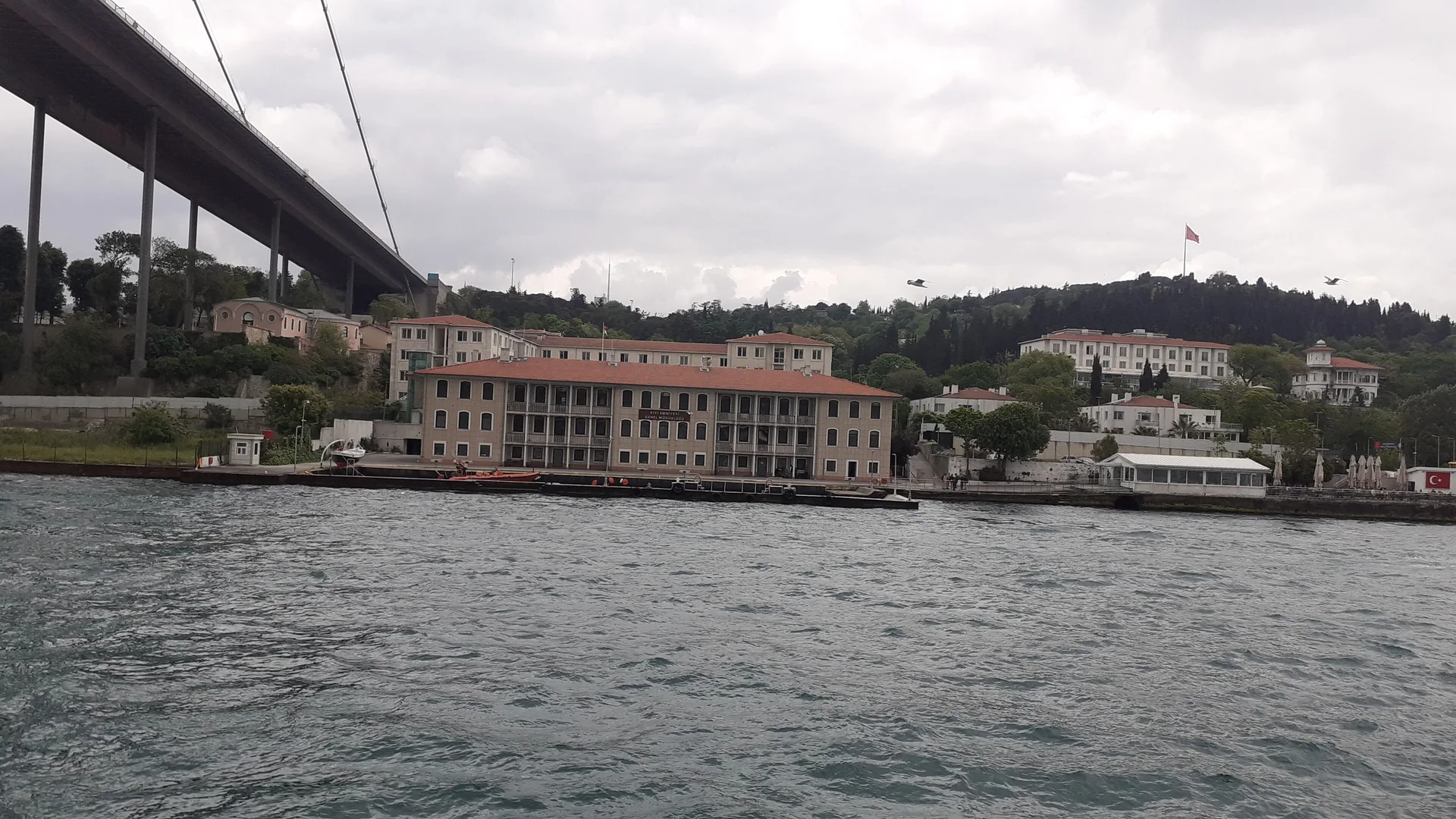 Boğaz kıyısında köprü altında kiremit çatılı büyük bina ve yeşil tepeler, önde dalgalı deniz