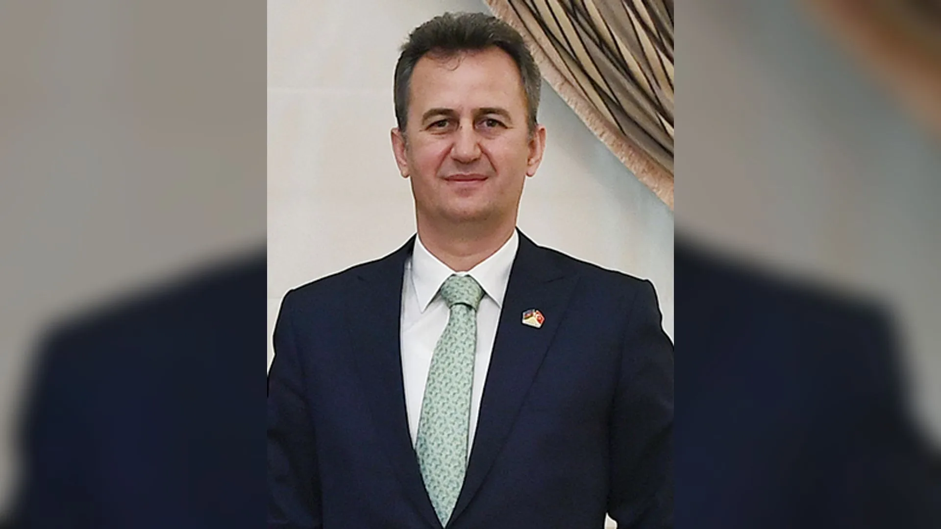 Lacivert takım elbiseli, açık yeşil kravatlı, yakasında rozet olan kısa saçlı bir adam