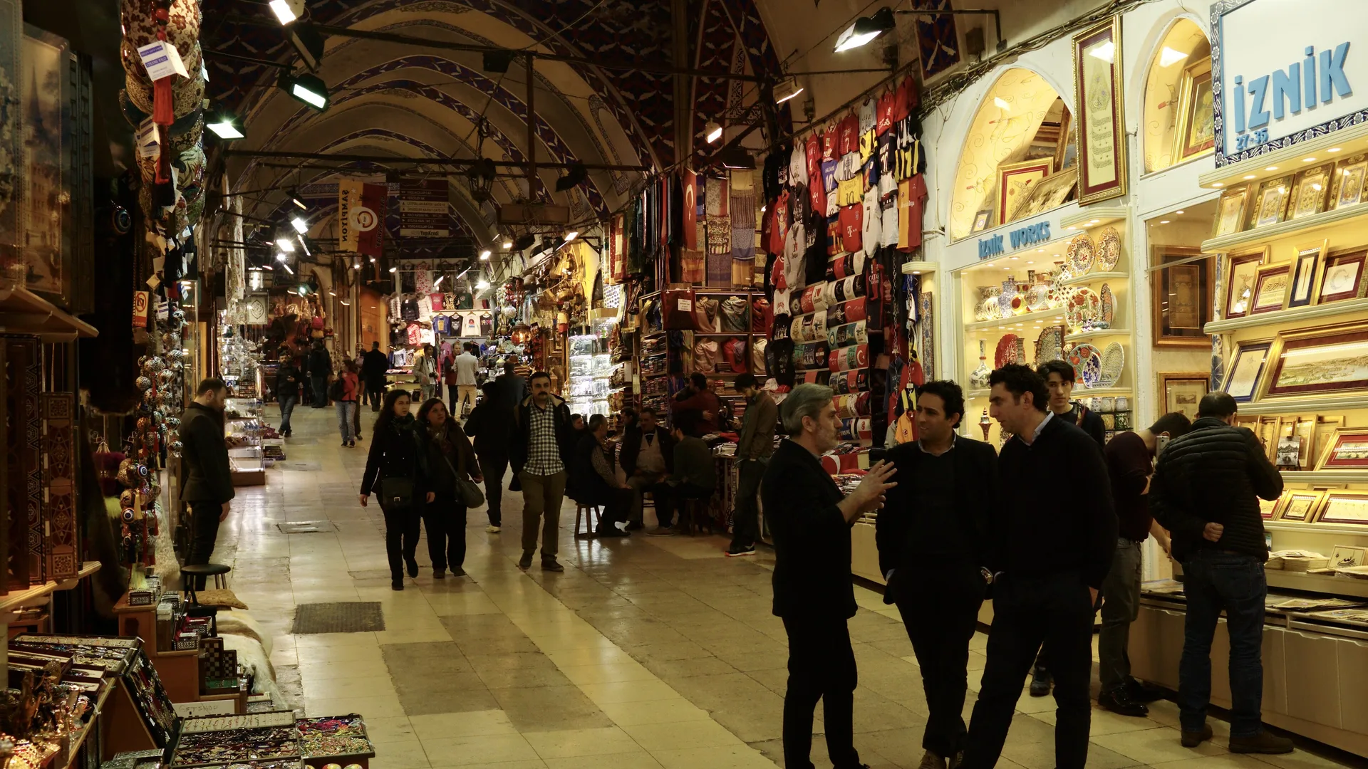Kemerli tavanlı, ışıklı bir çarşı koridoru; dükkânlar ve önde sohbet eden adamlar